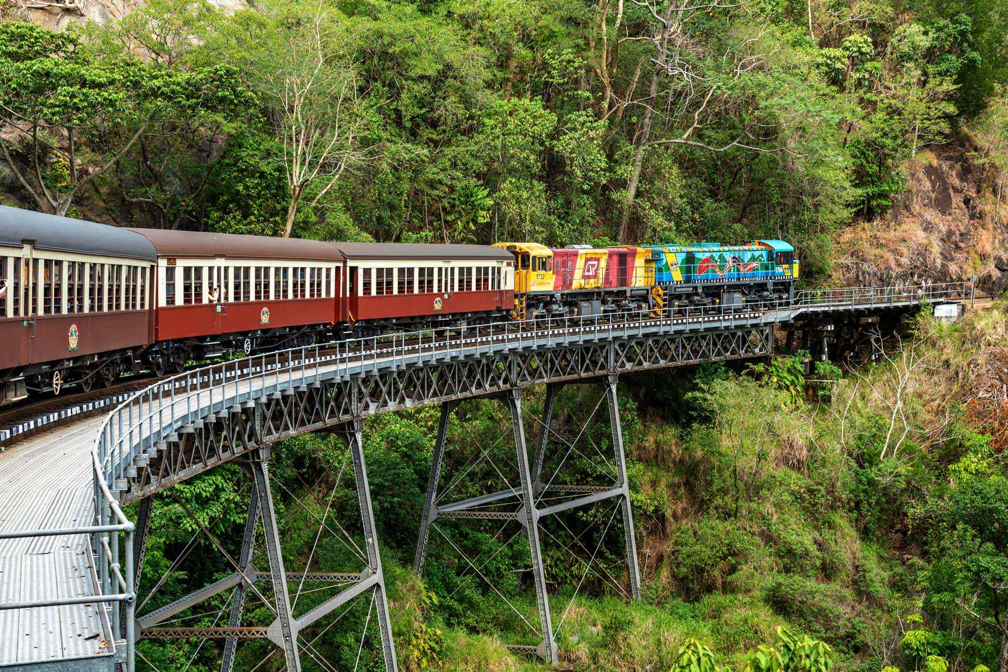 A colorful diesel locomotive and vintage passenger train cars traverse a curved metal trestle bridge spanning above lush rainforest canopy. The train features vibrant red and cream passenger carriages with a multi-colored diesel engine leading the consist across the dramatic iron lattice bridge structure.