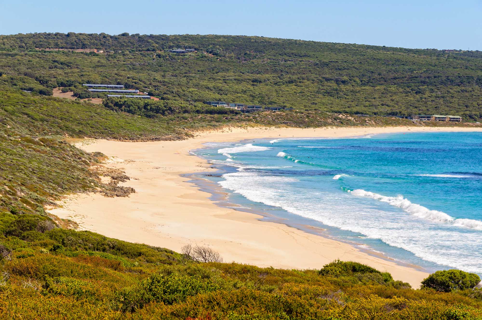 A stunning panoramic view of Smiths Beach near Yallingup, Western Australia, featuring golden sand, turquoise waters with rolling white-capped waves, and lush green coastal vegetation covering the hillside above. Scattered residential properties are visible nestled among the native bushland overlooking this pristine beach destination.