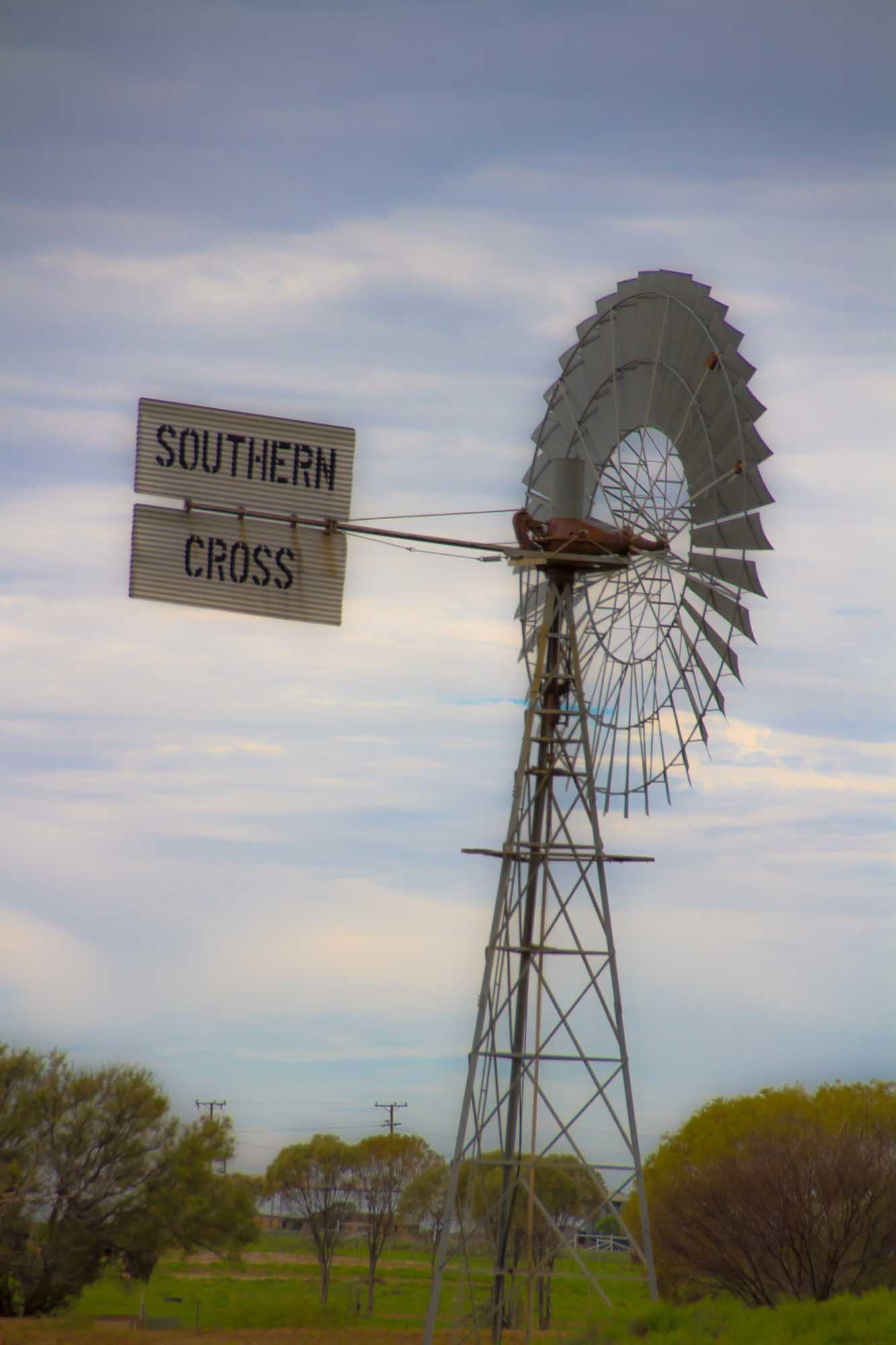 A historic American-style windmill with the 'Southern Cross' nameplate stands prominently in the arid Australian outback landscape near Longreach, Queensland. The tall metal structure features a distinctive multi-blade windmill wheel set against a partly cloudy sky, with sparse vegetation and utility poles visible in the surrounding flat terrain.