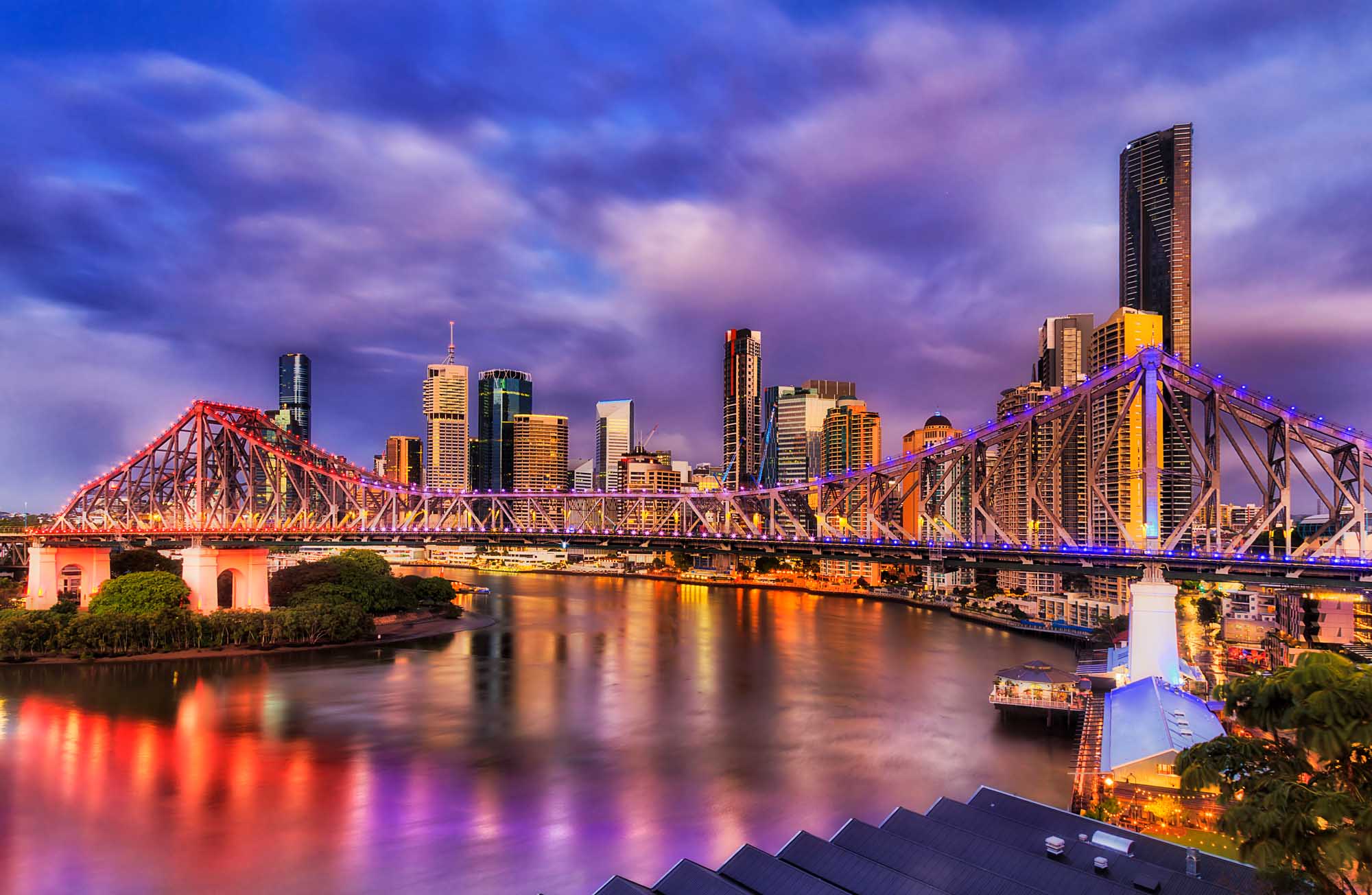 The iconic Story Bridge spans the Brisbane River in Queensland's central business district, illuminated in vibrant red and purple lights during the twilight hour. The bridge's distinctive steel lattice structure is beautifully reflected in the calm water below, with the modern skyline of Brisbane's towers glowing warmly in the background.