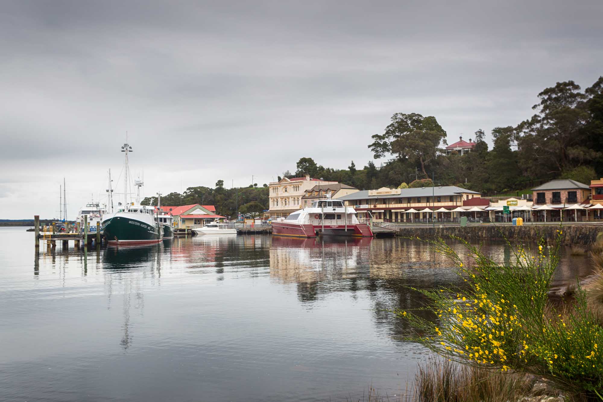A picturesque view of Strahan, a historic fishing town on Tasmania's west coast, featuring moored vessels including fishing boats and tourist cruisers reflected in calm waters, with heritage buildings and tree-lined shores visible in the background.