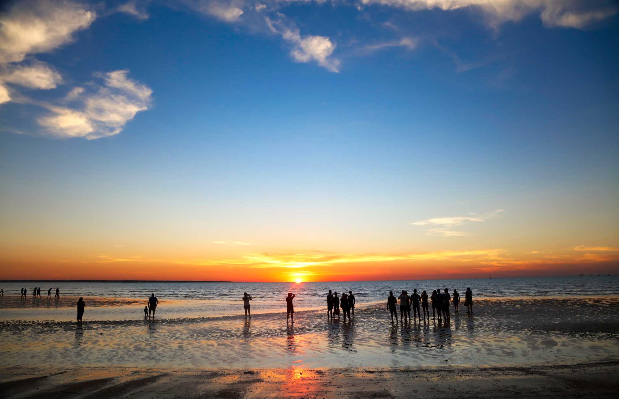 A scenic sunset view at Mindil Beach in Darwin, with silhouetted crowds of people wading in the shallow waters and standing on the beach, watching the golden sun descend over the ocean horizon under a partly cloudy sky.