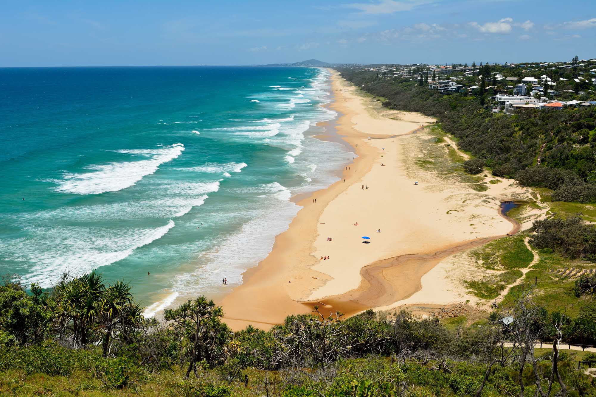 An aerial view of a pristine golden sandy beach stretching along the Queensland coastline, with turquoise ocean waves, lush green hinterland vegetation, and residential properties dotting the clifftop. The beach features a natural sandbar formation and scattered beachgoers enjoying the clear summer day.