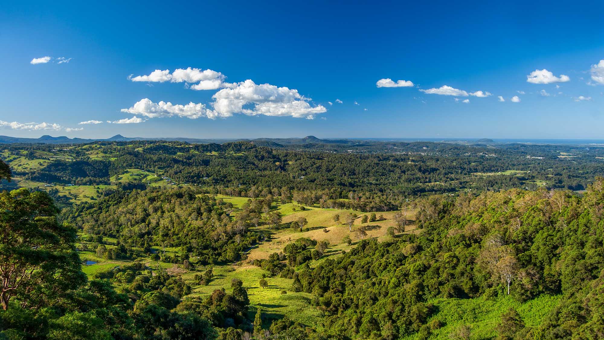 A sweeping aerial view of the lush Sunshine Coast Hinterland in Queensland, Australia, featuring rolling green hills, scattered forests, pastoral farmland, and distant mountains under a brilliant blue sky with white cumulus clouds.