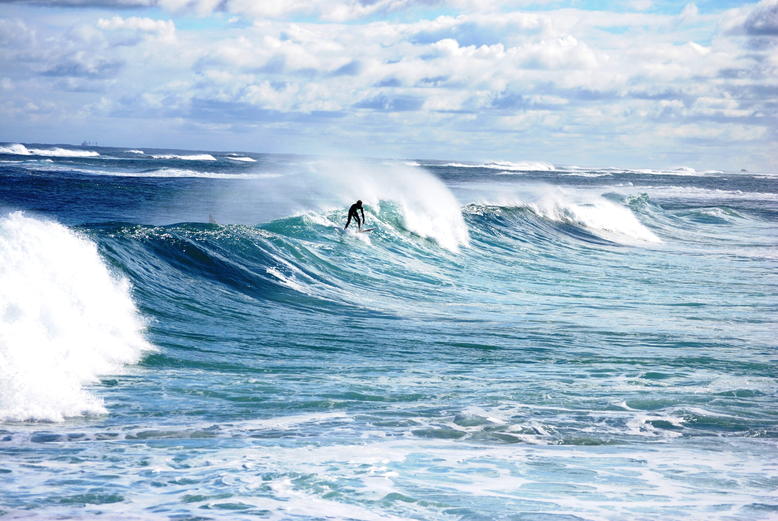 A skilled surfer in a wetsuit rides a powerful turquoise and white wave beneath a partly cloudy blue sky. Multiple waves can be seen in the background, indicating excellent surfing conditions with consistent swell.