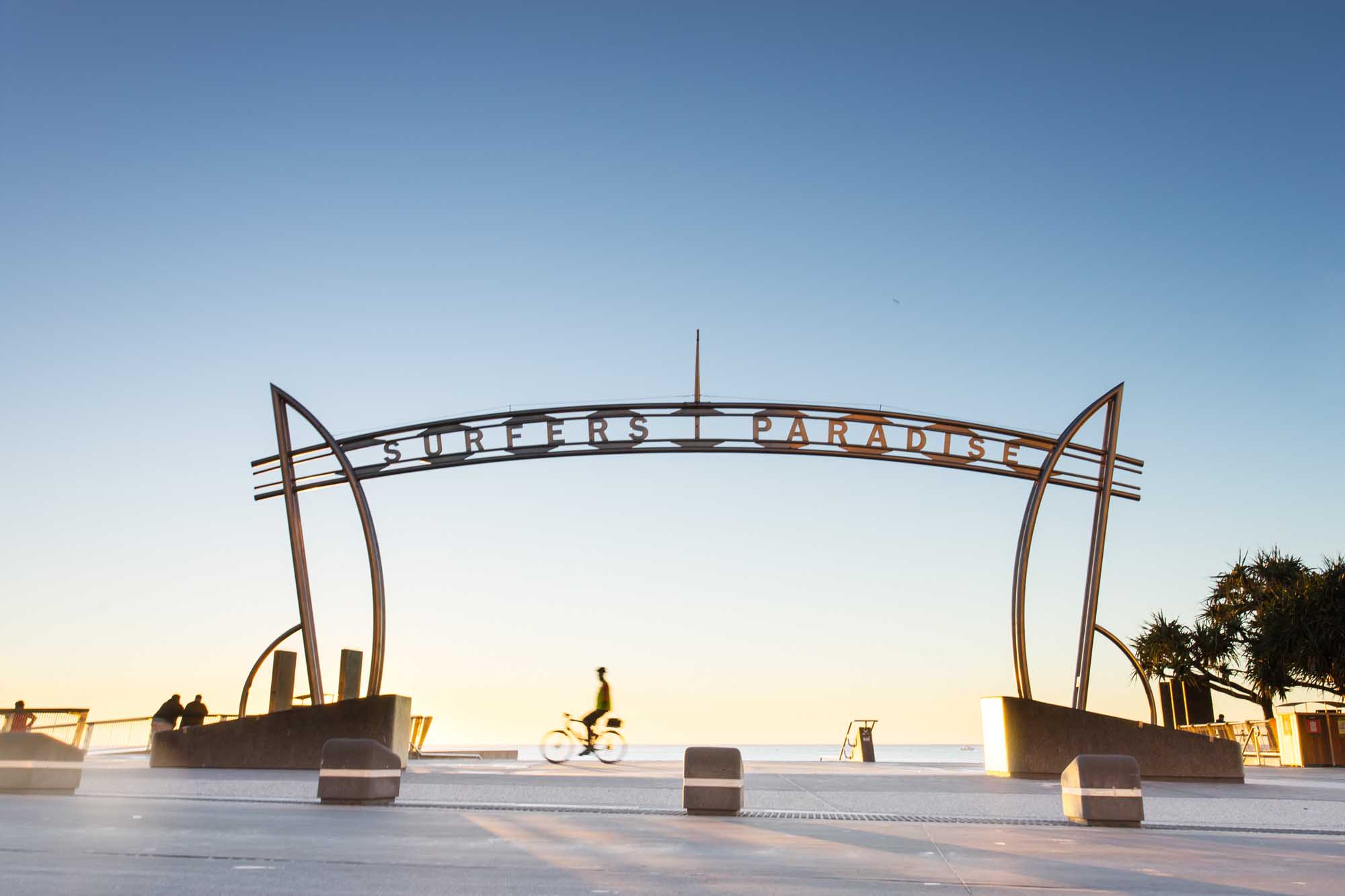 The iconic 'Surfers Paradise' archway sign stands prominently at sunrise on Queensland's Gold Coast in Australia. The sculptural steel structure frames the beach and ocean beyond, with a cyclist passing through and golden light illuminating the seaside promenade in the early morning hours.