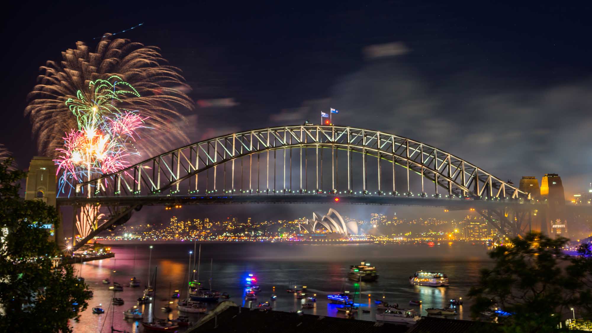 A spectacular nighttime view of Sydney's iconic Harbour Bridge illuminated during New Year's Eve celebrations, with vibrant fireworks exploding above the bridge and the illuminated Sydney Opera House visible below. Colourful boats dot the harbour waters as the city celebrates with spectacular pyrotechnics reflecting across the water.