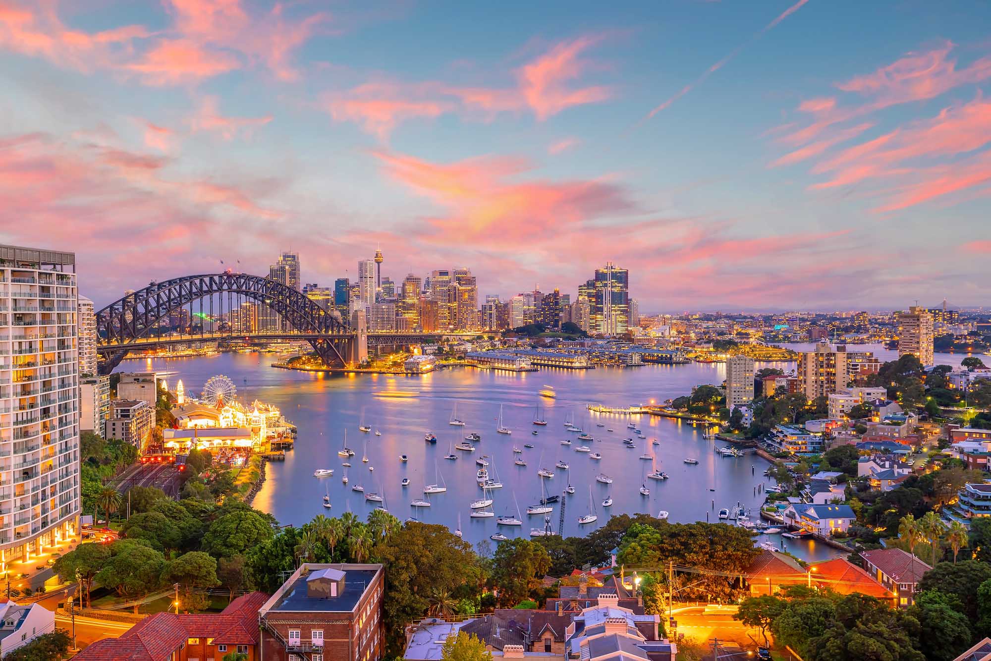 A stunning twilight view of Sydney Harbour featuring the iconic Sydney Harbour Bridge, illuminated skyline, moored sailboats, and the Sydney Opera House in the distance. The image captures the vibrant evening atmosphere of Australia's most recognizable waterfront, with warm golden lights reflecting across the calm water and colorful pink and blue sky.