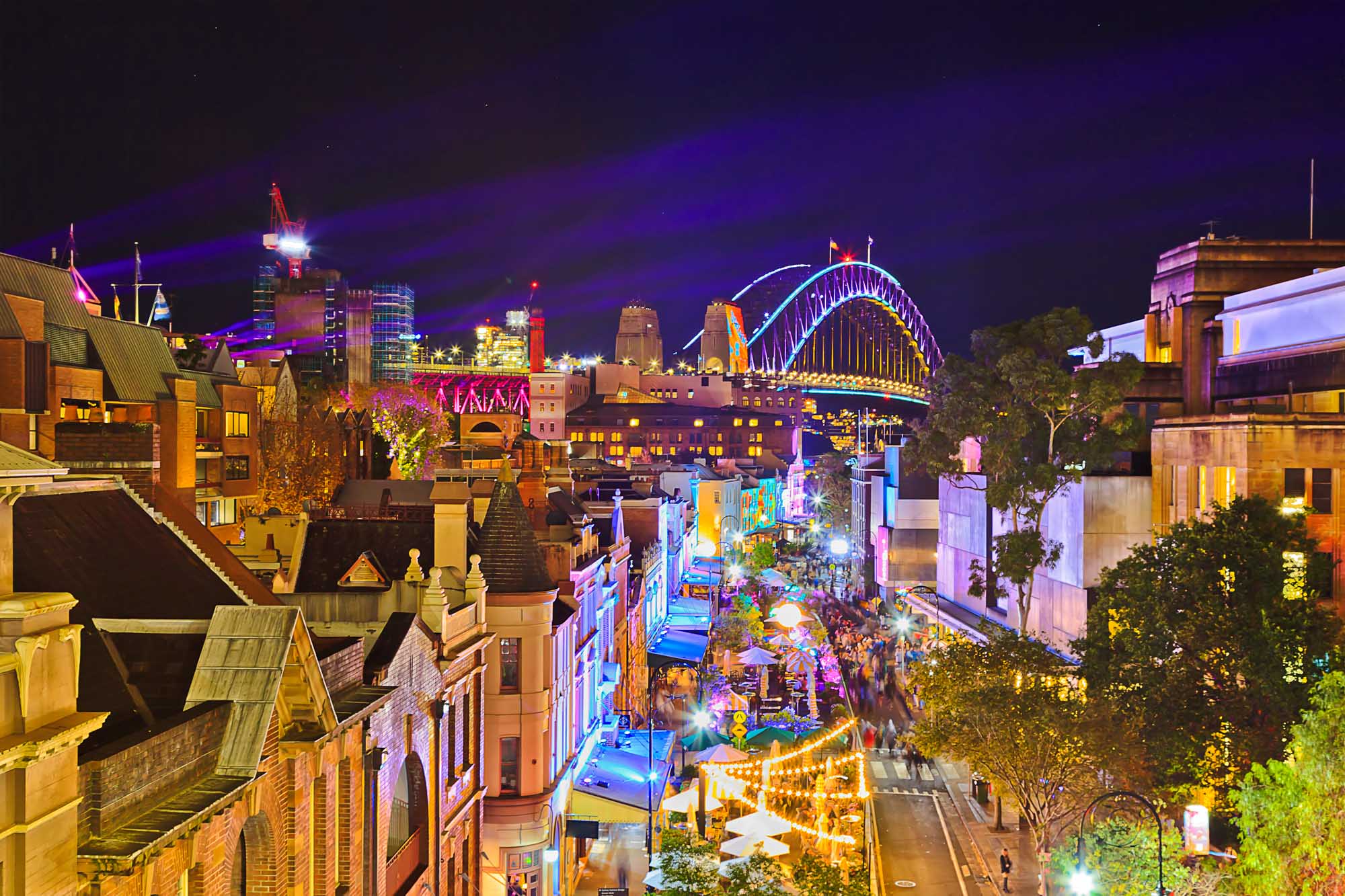A vibrant nighttime cityscape of Sydney, Australia during the Vivid Sydney light show, featuring the iconic Sydney Harbour Bridge illuminated in blue and gold, the Opera House, and surrounding buildings bathed in colorful projected lighting. The pedestrian mall below is bustling with visitors enjoying the festival's interactive light installations and street performances.