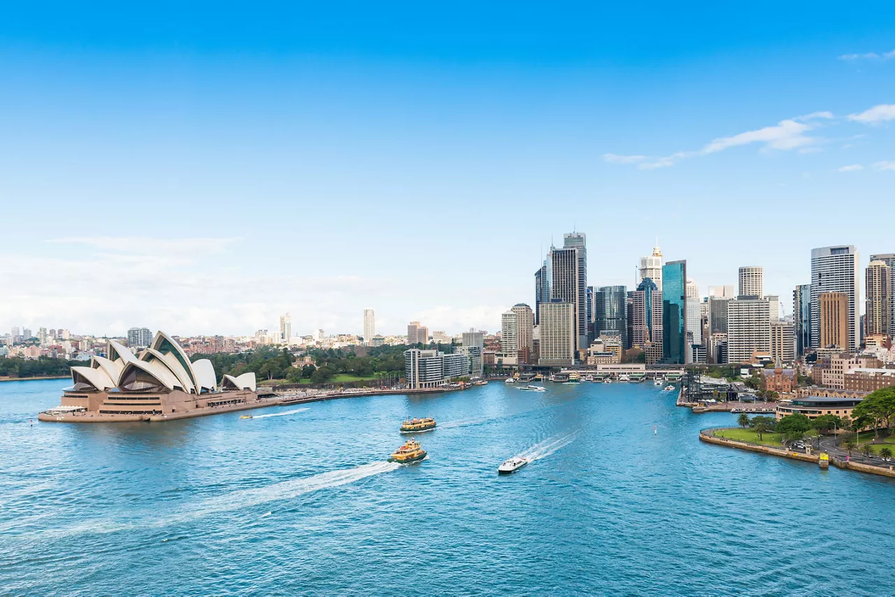 A stunning panoramic view of Sydney, Australia's iconic harbour featuring the distinctive white shells of the Opera House on the left, bustling with ferries and water traffic, with the gleaming modern skyline of downtown Sydney rising behind the sparkling blue waters of Circular Quay.