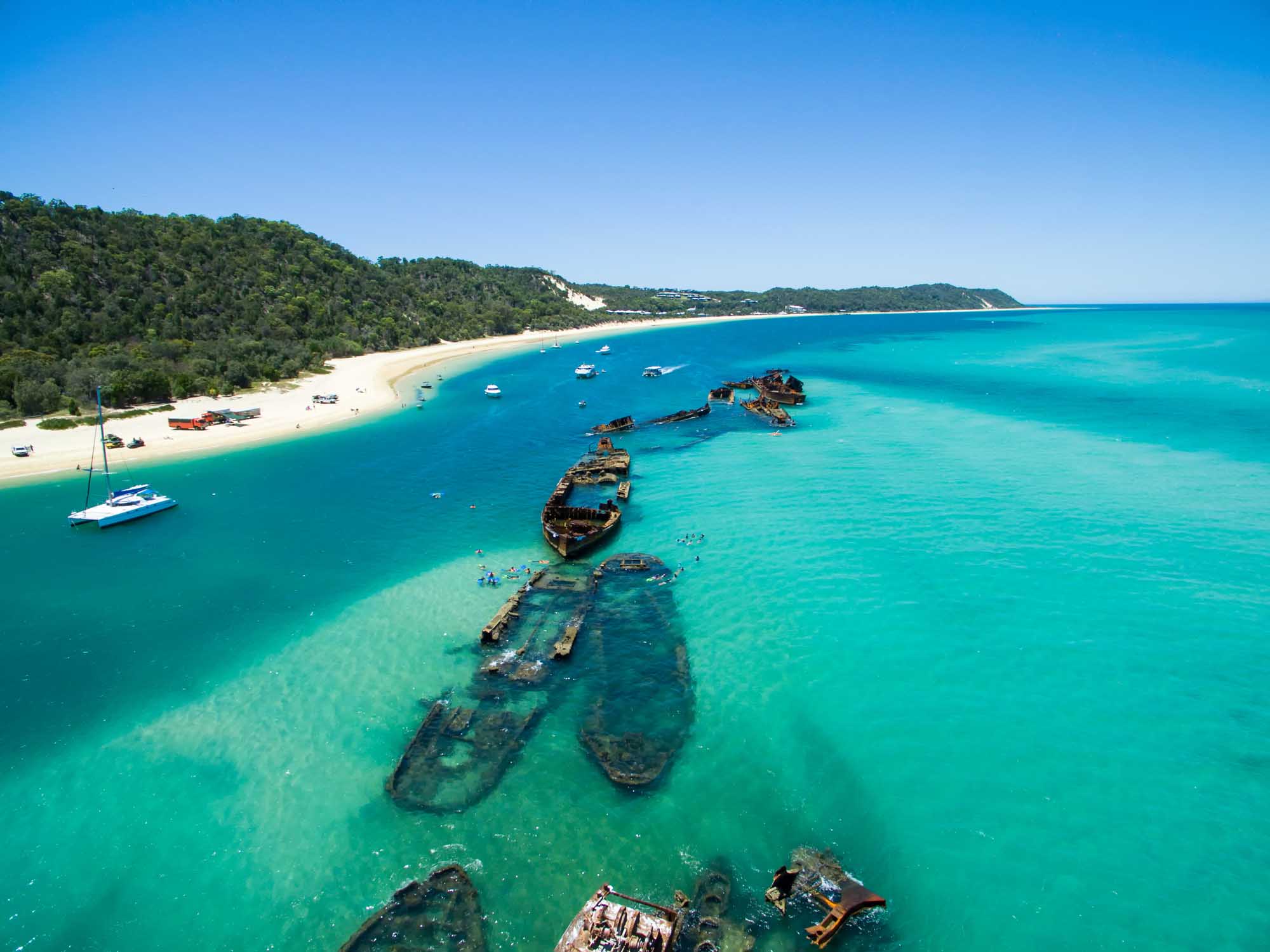 An aerial view of the Tangalooma shipwrecks submerged in the turquoise waters off Moreton Island, Queensland, Australia. The image captures multiple sunken vessels in shallow water with a pristine sandy beach and forested coastline in the background, along with moored boats and swimmers exploring the historic wreck site.