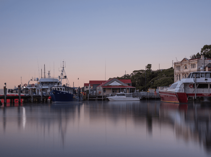 A serene waterfront scene at dawn featuring several moored fishing and commercial vessels reflected in calm harbor waters, with historic waterfront buildings and tree-lined hills in the background under a clear blue sky.