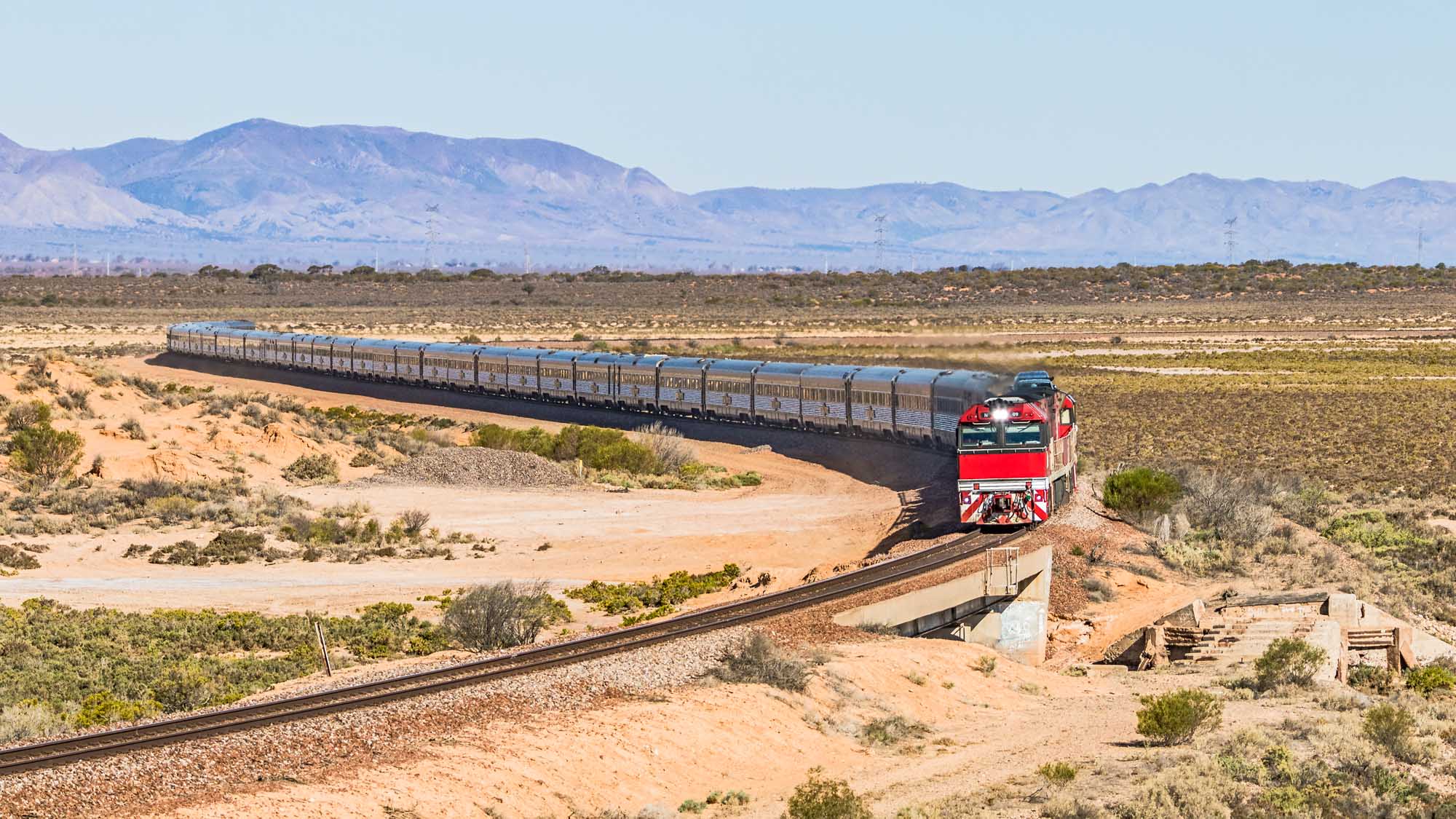 A long-distance luxury passenger train with distinctive red engines curves through the arid South Australian outback, with the dramatic Flinders Ranges mountains visible on the horizon. The train travels through desert terrain dotted with sparse vegetation, heading north from Adelaide toward Darwin.