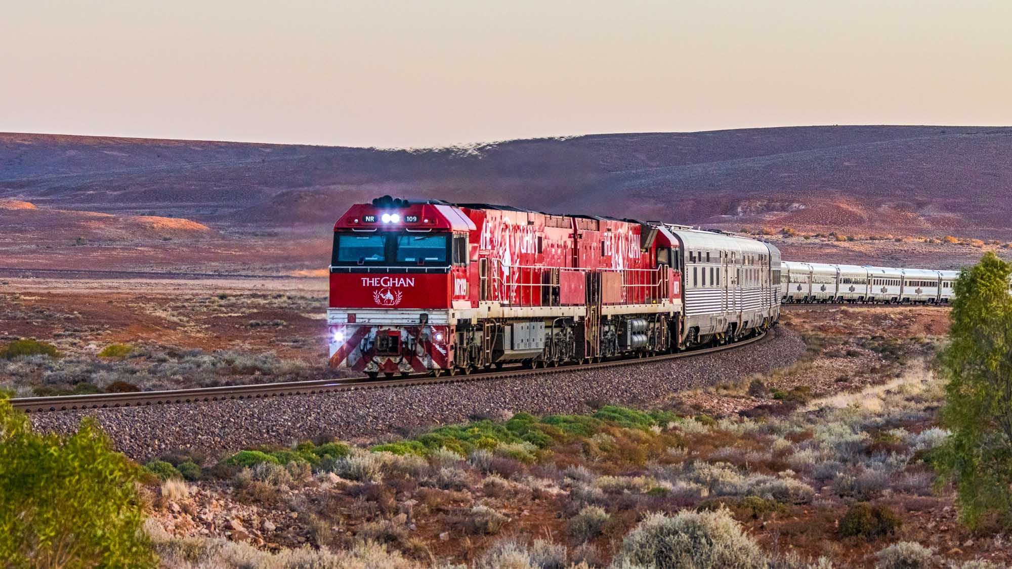 Australia's legendary Ghan luxury tourist train, operated by Journey Beyond Rail, travels through the remote South Australian outback near Pimba at sunset. The distinctive red and cream consist, pulled by two specially painted GE locomotives bearing the iconic camel logo, rounds dramatic curves against a striking mesa backdrop in the Australian interior.