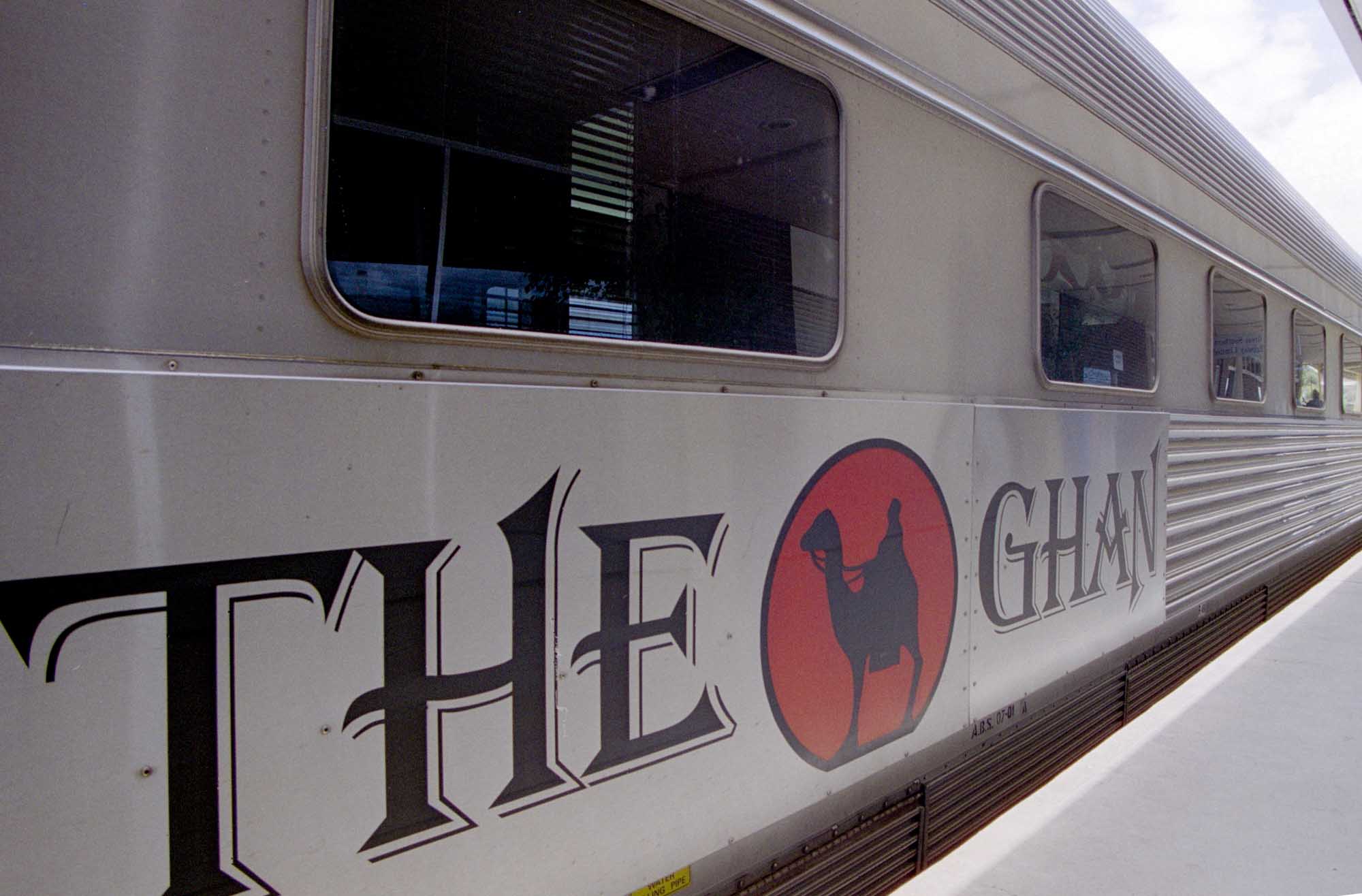 A side view of The Ghan, Australia's famous long-distance passenger railway train, displaying the distinctive branding with stylized lettering and red circular logo on the silver and grey exterior. The image shows multiple windows along the carriage and corrugated metal paneling typical of this iconic Australian train service.