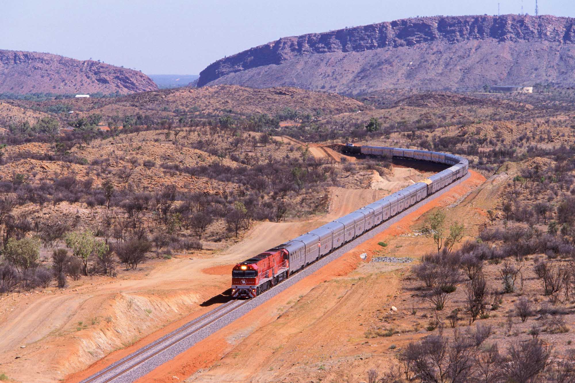 A long passenger train with red locomotive winds through the arid Australian outback landscape near Alice Springs, with the MacDonnell Ranges visible in the background. The iconic Ghan transcontinental railway is depicted traveling through Australia's Red Centre on its inaugural journey from Adelaide to Darwin.