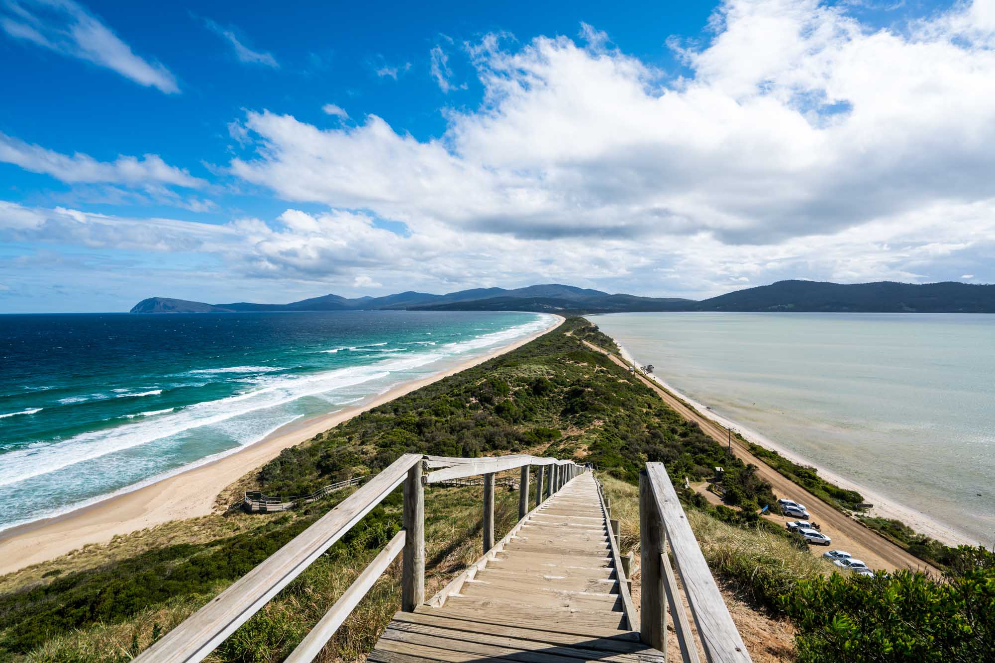 A dramatic aerial view of The Neck, a narrow isthmus on Bruny Island, featuring a wooden boardwalk descending toward pristine white sand beaches flanked by turquoise ocean waters on one side and calm bay waters on the other, with lush green vegetation covering the peninsula and mountains visible across the water under a partly cloudy sky.