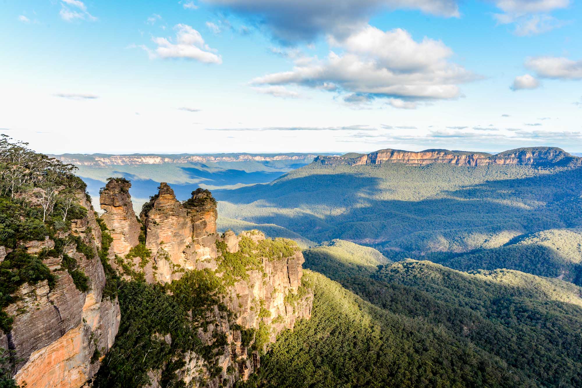 A stunning panoramic view of the iconic Three Sisters rock formation in the Blue Mountains of New South Wales, Australia, with dramatic sandstone peaks rising above lush eucalyptus forest and distant mountain ridges under a partly cloudy sky.