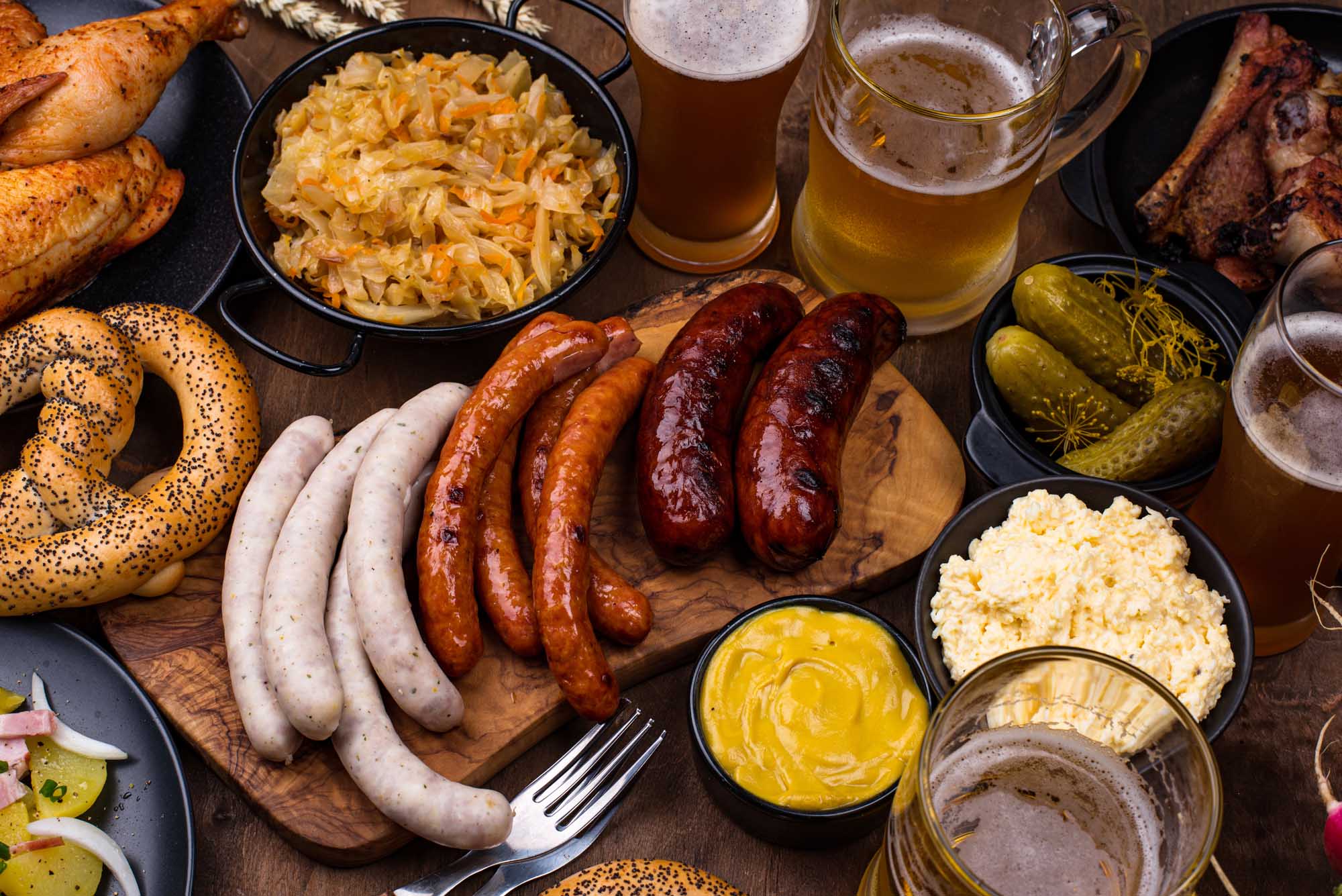 An authentic Oktoberfest spread featuring an assortment of traditional German dishes including various sausages, soft pretzels, roasted chicken, ribs, stewed cabbage, potato salad, pickles, and mustard, accompanied by multiple glasses of fresh beer. The feast is artfully arranged on wooden boards and in traditional serving vessels, capturing the festive spirit of Bavarian celebration.