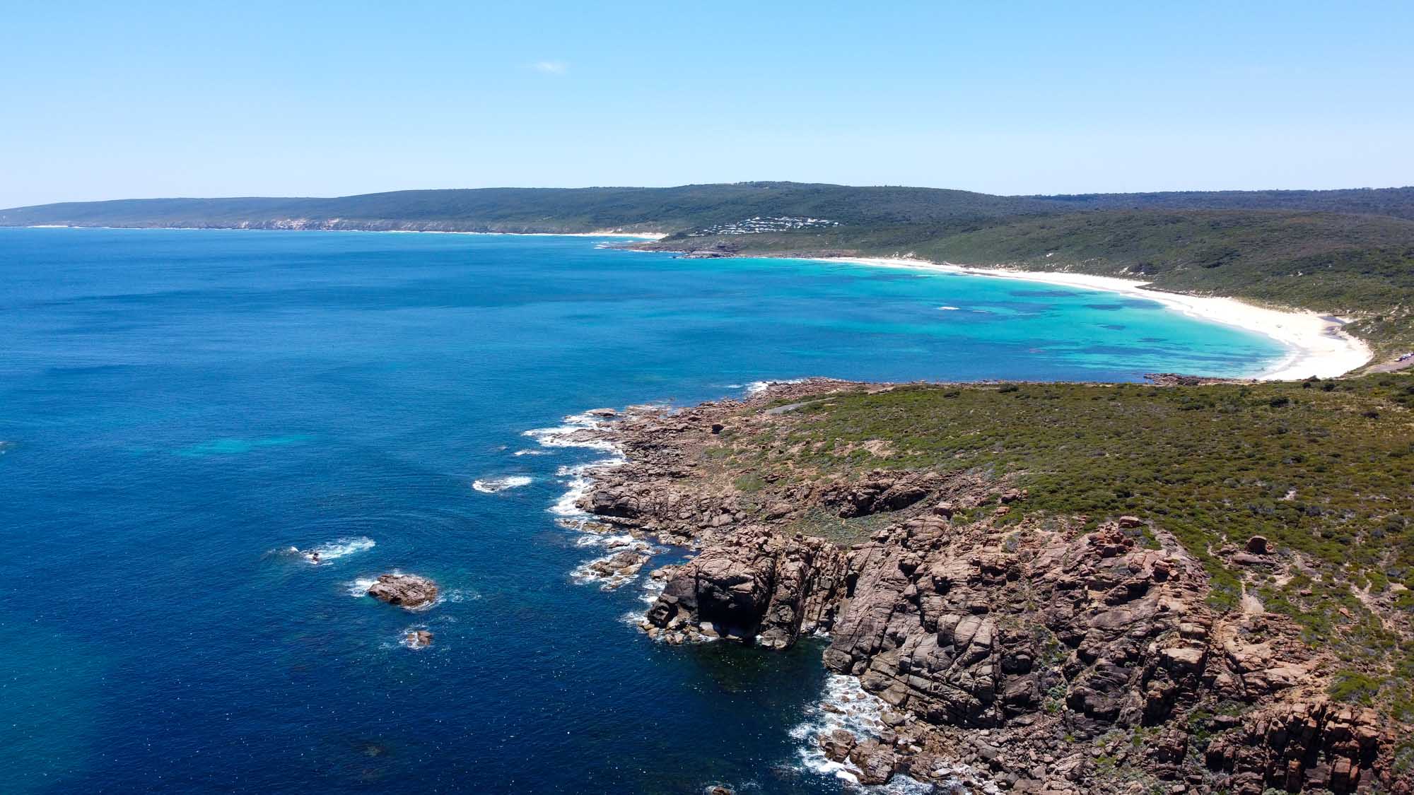 A stunning aerial view of a crescent-shaped white sand beach nestled against turquoise waters and rugged rocky headlands along the southwestern Australian coastline. The dramatic granite cliffs and lush vegetation frame a pristine, secluded cove with crystal-clear shallow waters that transition from emerald to deep blue.
