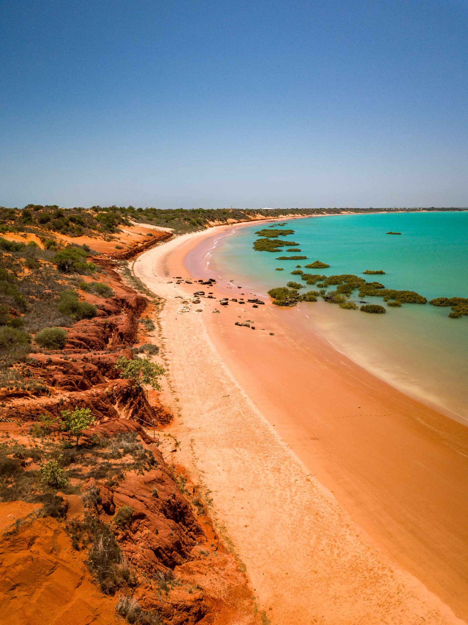 A stunning aerial view of Roebuck Bay in Broome, Western Australia, showcasing pristine turquoise waters, a curved white sand beach, and distinctive red ochre cliffs dotted with native vegetation. Mangrove formations dot the shallow waters, creating a dramatic contrast with the vibrant blue sea and clear sky.