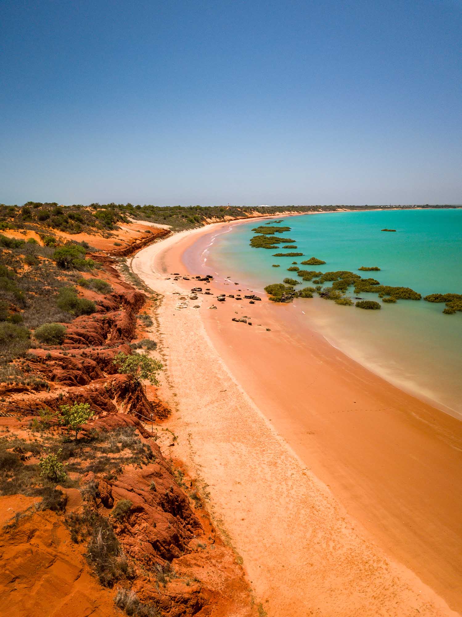 A stunning aerial view of Roebuck Bay in Broome, Western Australia, showcasing pristine turquoise waters, a curved white sand beach, and distinctive red ochre cliffs dotted with native vegetation. Mangrove formations dot the shallow waters, creating a dramatic contrast with the vibrant blue sea and clear sky.