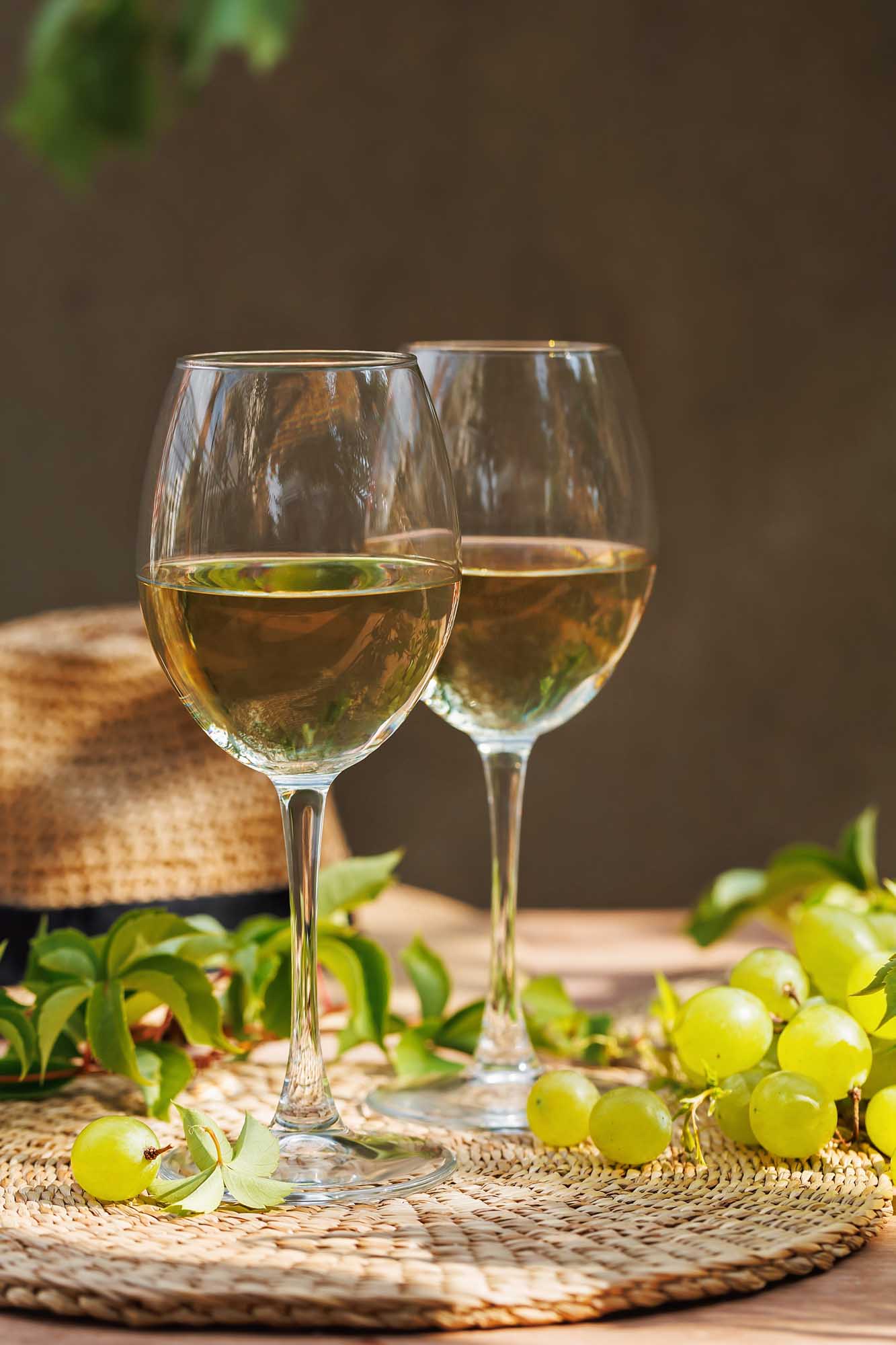 Two glasses of crisp white wine are displayed on a woven placemat alongside fresh green grapes, grapevine leaves, and a rustic straw hat, evoking the classic wine culture of Tuscany. The warm, natural lighting and earthy tones create an inviting scene ideal for wine tourism and culinary travel content.