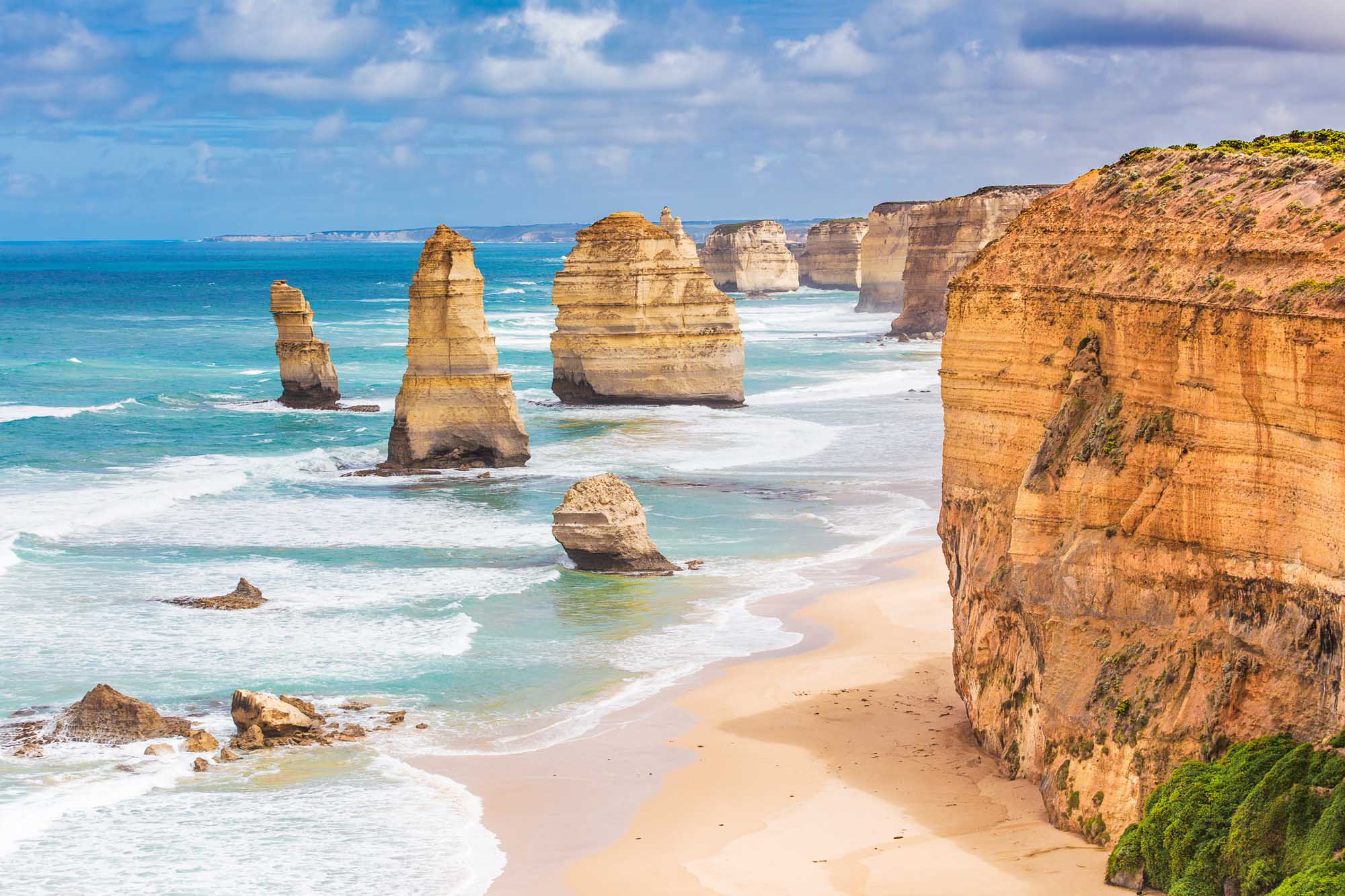 Dramatic limestone sea stacks rise majestically from the turquoise waters and sandy beach of the Twelve Apostles, an iconic natural formation along Australia's Great Ocean Road. Weathered golden cliffs frame the scene under a partly cloudy sky, showcasing the raw coastal beauty of Victoria's rugged shoreline.