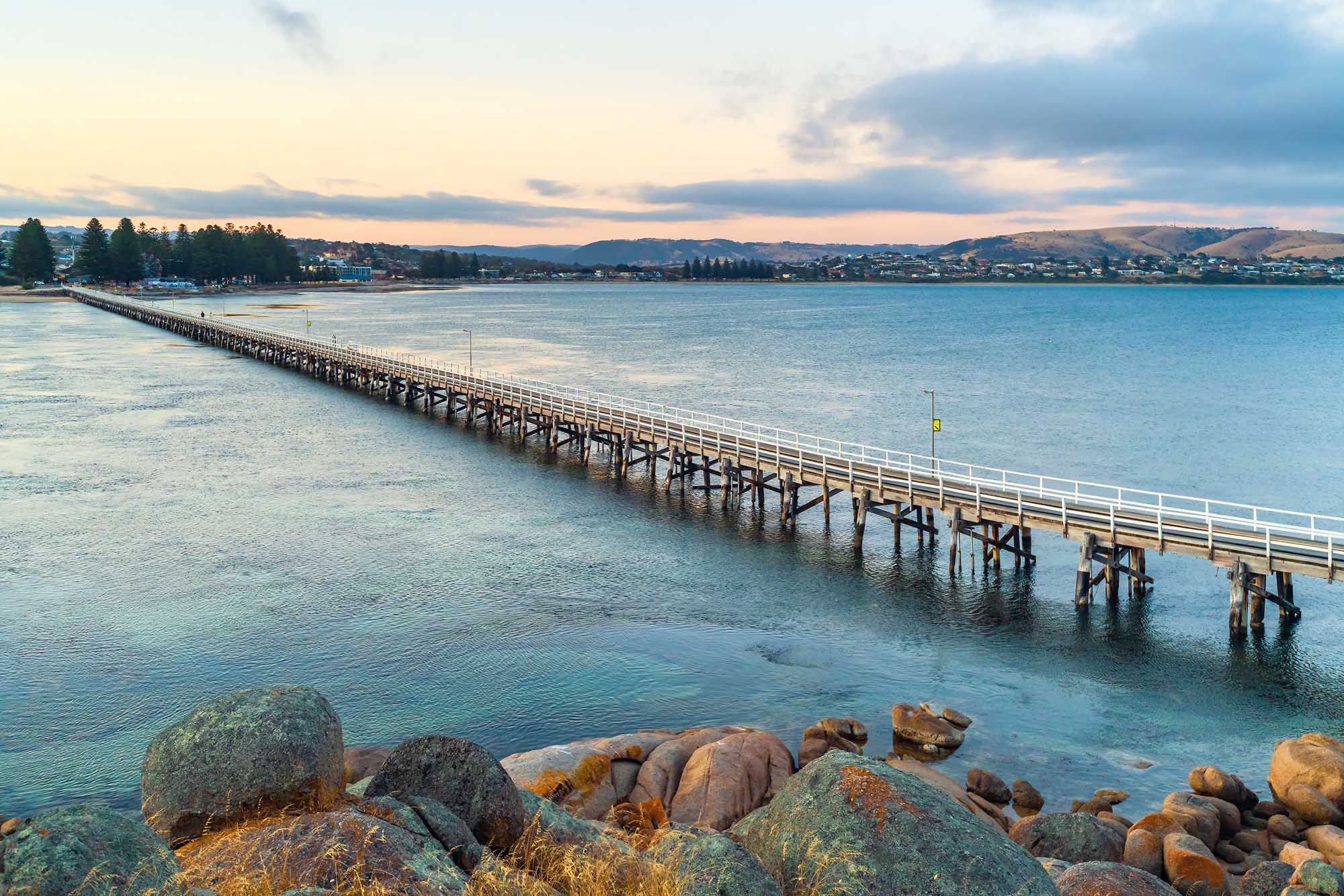 A scenic photograph of the iconic Victor Harbor Causeway stretching into calm turquoise waters at sunset, with granite boulders in the foreground and the coastal town of Victor Harbor visible along the shoreline beneath rolling hills.