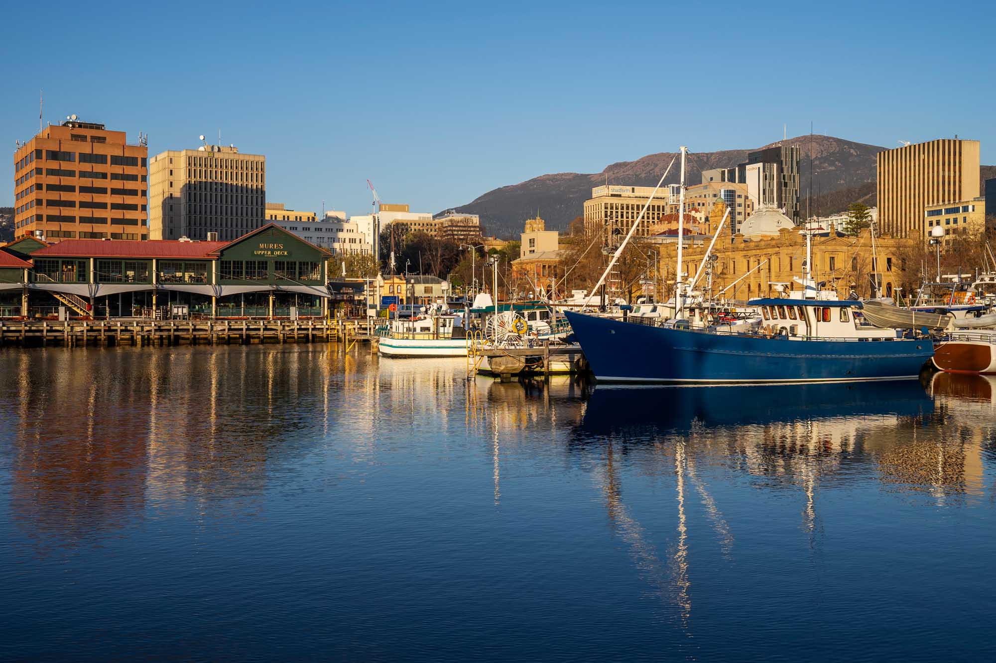 A scenic harbor view of Victoria Dock in Hobart, Tasmania, featuring moored fishing vessels and leisure boats reflected in calm blue waters at sunrise, with the distinctive Mount Wellington rising in the background and historic waterfront buildings lining the dock.