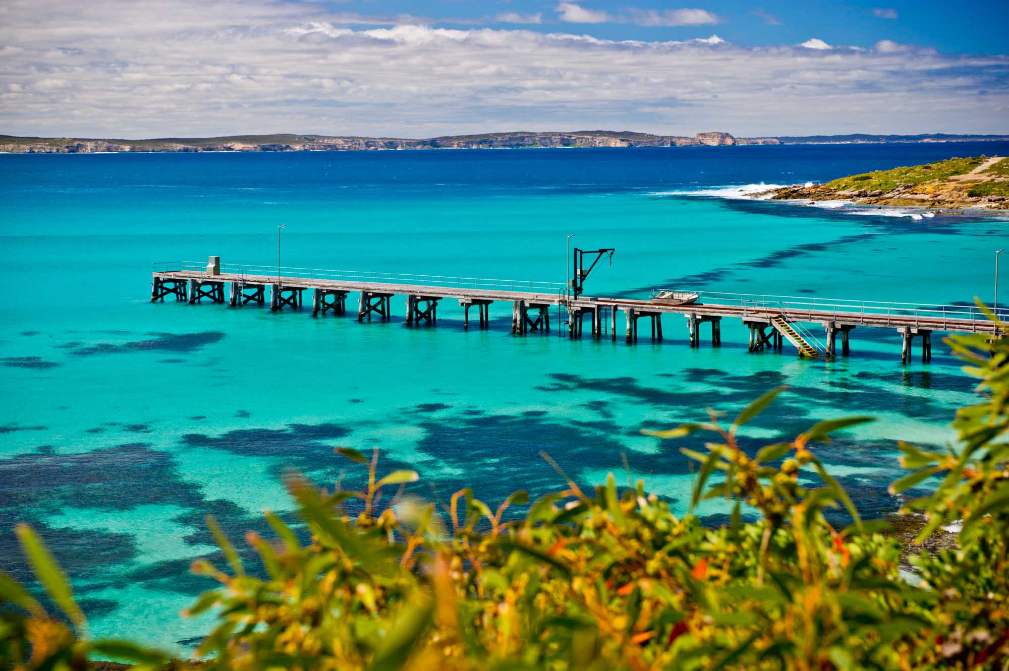 A stunning aerial view of Vivonne Bay on Kangaroo Island featuring a long wooden pier extending into crystalline turquoise waters, with dramatic coastal cliffs visible across the bay under a partly cloudy sky.