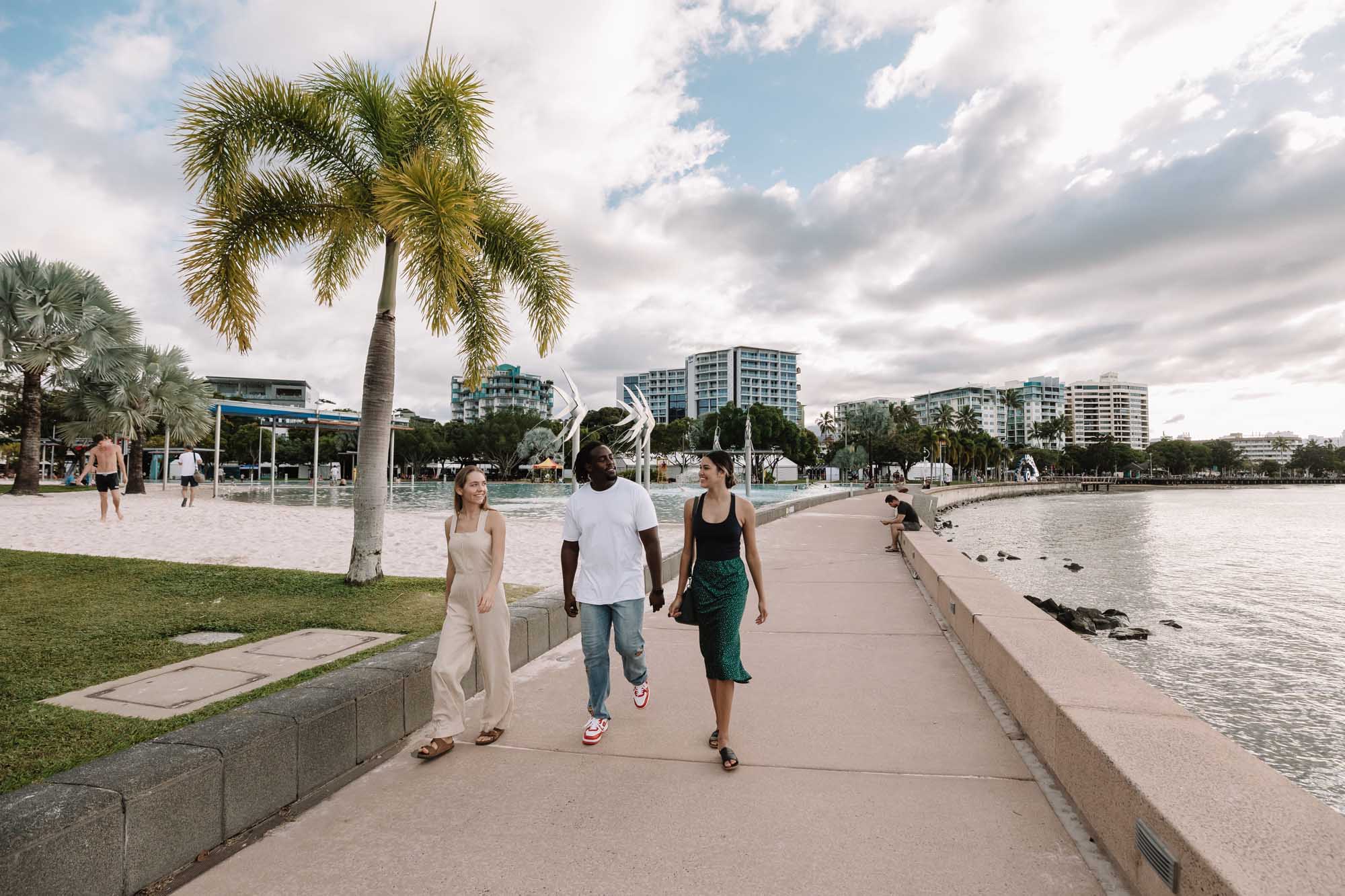 Three people jogging and walking along a scenic waterfront promenade at Cairns Esplanade Lagoon, with sandy beach, palm trees, and modern high-rise buildings in the background. The image captures a tropical Australian destination with calm water, manicured lawns, and a vibrant community atmosphere under partly cloudy skies.