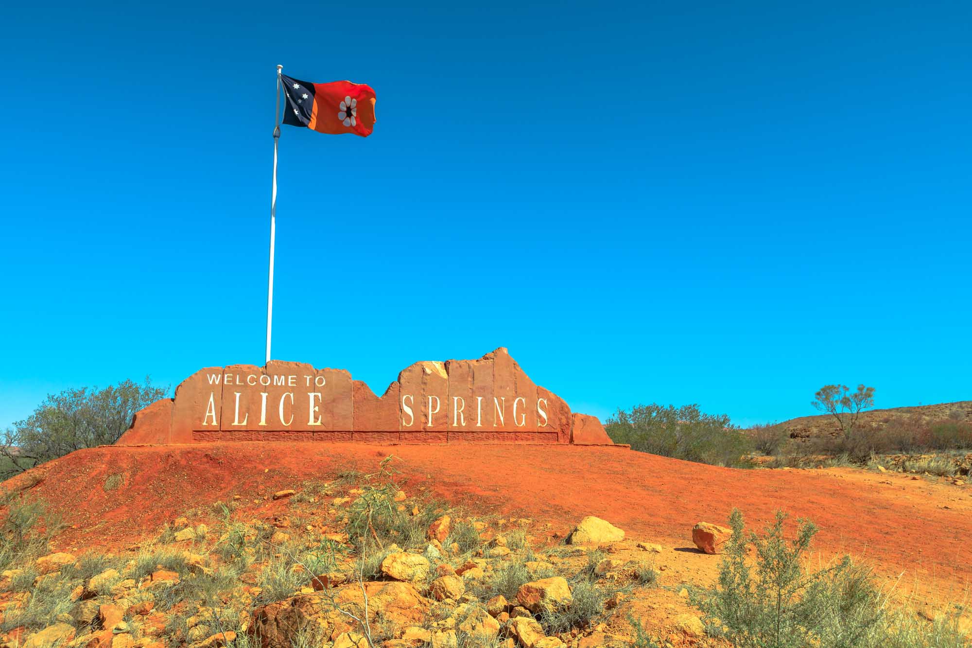 The iconic "Welcome to Alice Springs" sign carved into a red rock formation, flying the Northern Territory flag on a white flagpole against a clear blue sky. The arid outback landscape with sparse desert vegetation surrounds this popular tourism landmark in central Australia's Red Center.