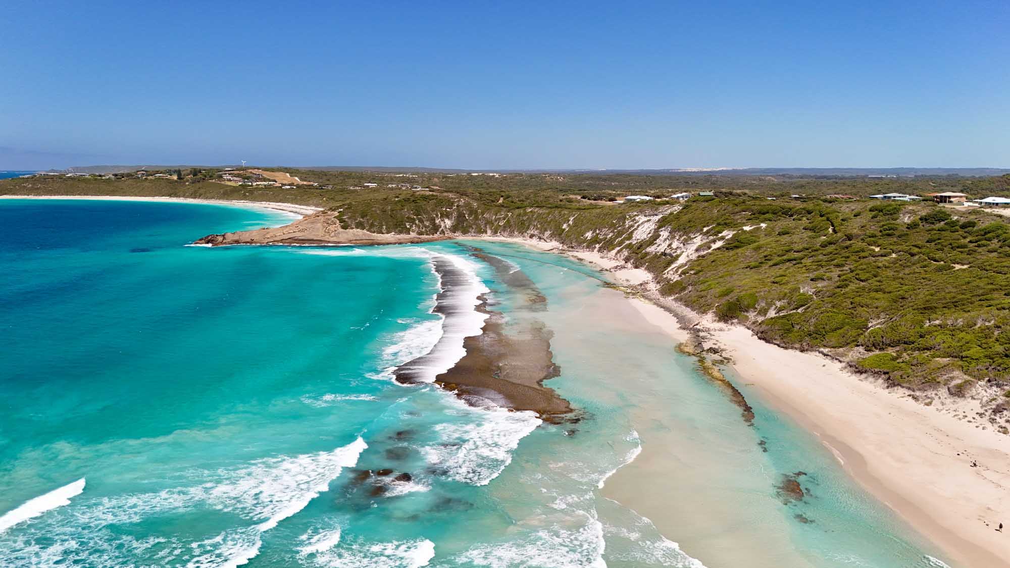 An aerial perspective of West Beach in Esperance, Western Australia, showcasing pristine white sandy shores bordered by dramatic rocky coastal formations and turquoise Southern Ocean waters. The image captures the dynamic contrast between the natural rugged coastline, calm beach coves, and the low-rise suburban development with residential neighborhoods, shops, and road networks characteristic of this regional coastal city.
