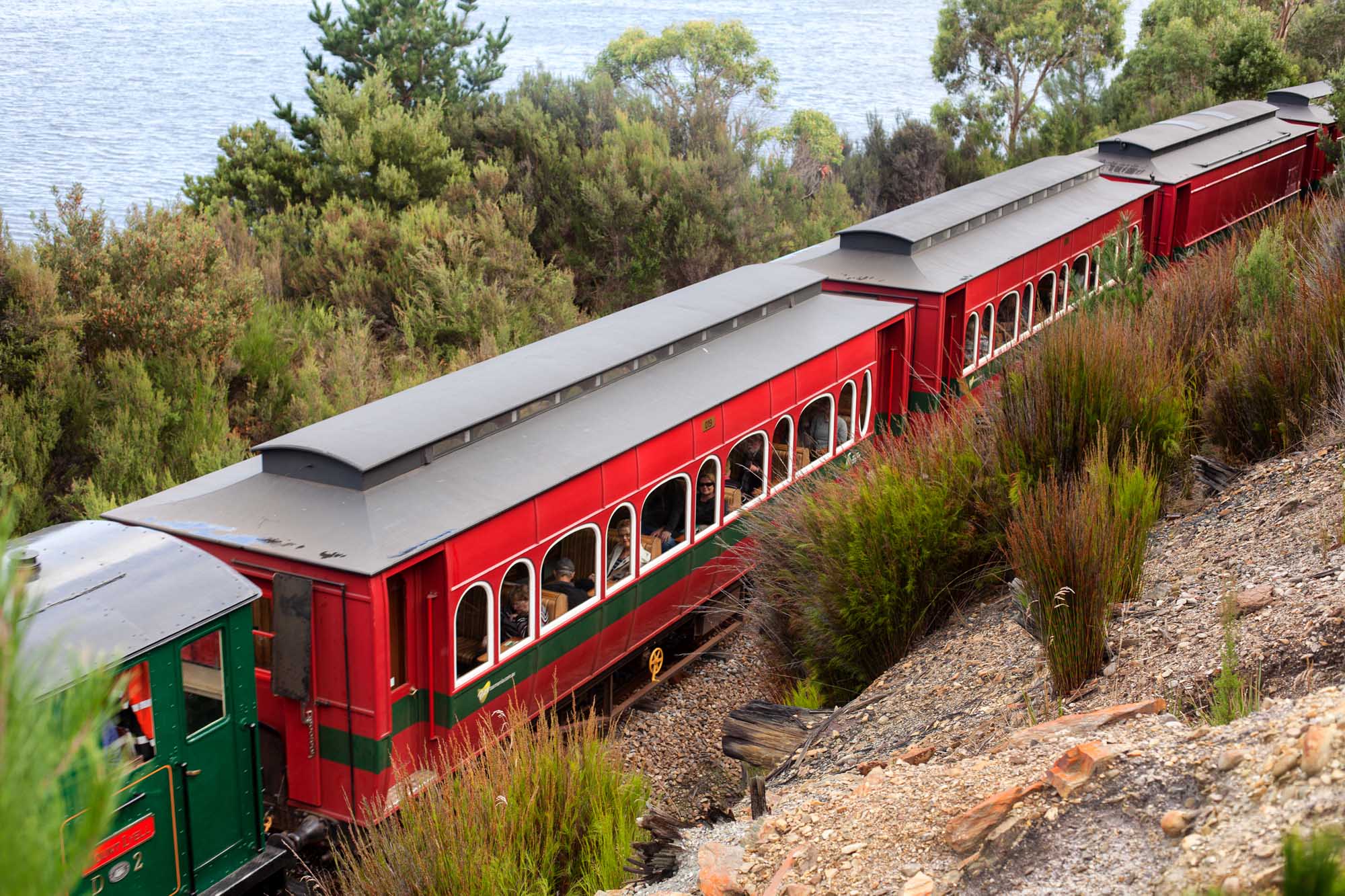 A red and green heritage locomotive with passenger carriages winds through mountainous rainforest terrain alongside a pristine body of water in Tasmania, Australia. The train operates on the Abt rack system, a specialized railway built to navigate the steep gradients of the West Coast Wilderness Railway route between Queenstown and Strahan.