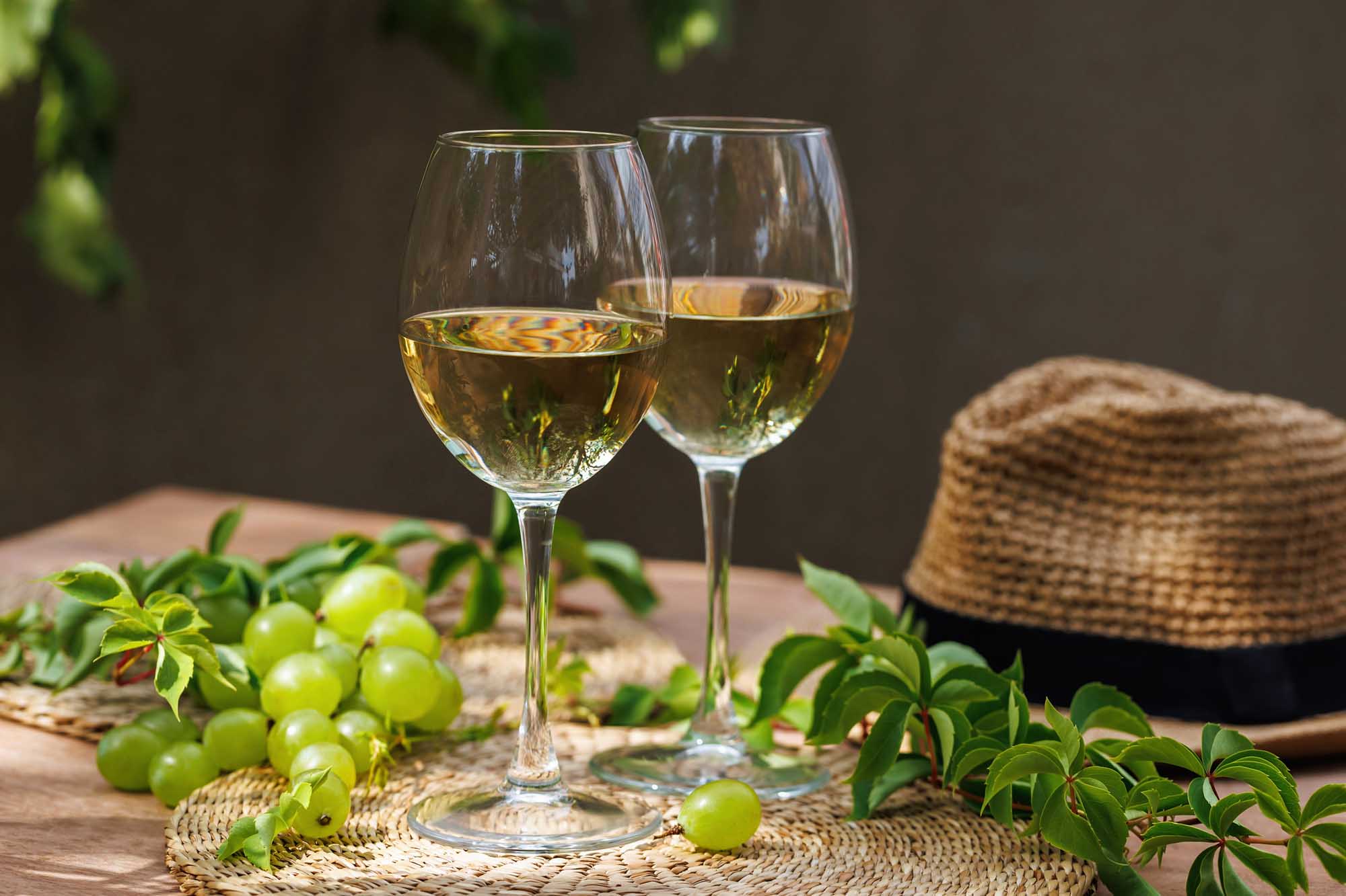 Two glasses of crisp white wine are elegantly arranged on a wooden table alongside fresh green grapes, green vine leaves, and a woven straw hat, creating a leisurely vineyard or wine country aesthetic with soft natural lighting.