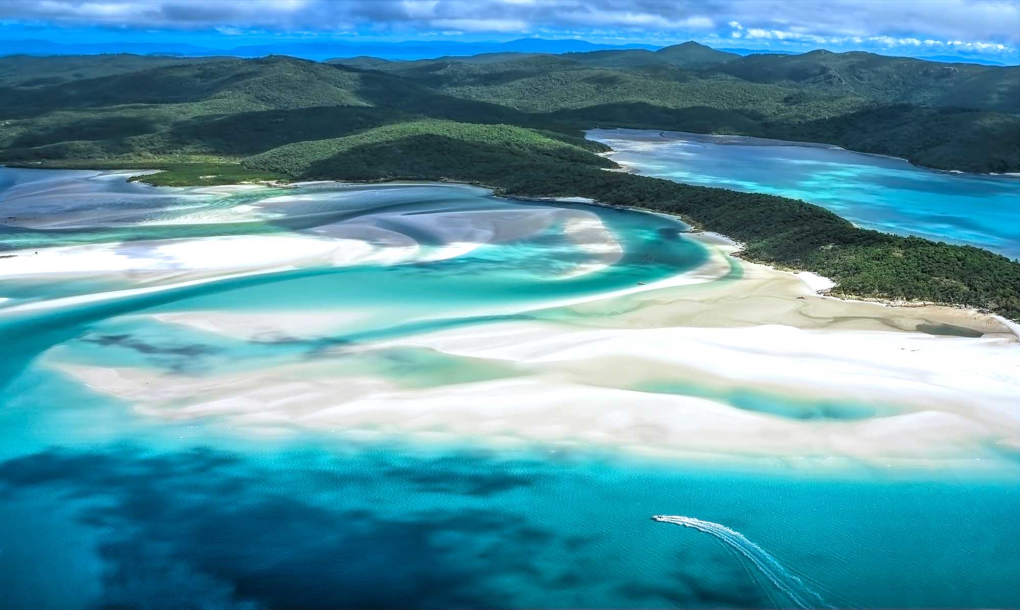 A stunning aerial photograph of Whitehaven Beach on Whitsunday Island, featuring pristine white silica sand, turquoise lagoons, and lush green hills. The image captures the iconic swirling sandbars and crystal-clear waters that make this Australian destination a world-renowned tropical paradise.