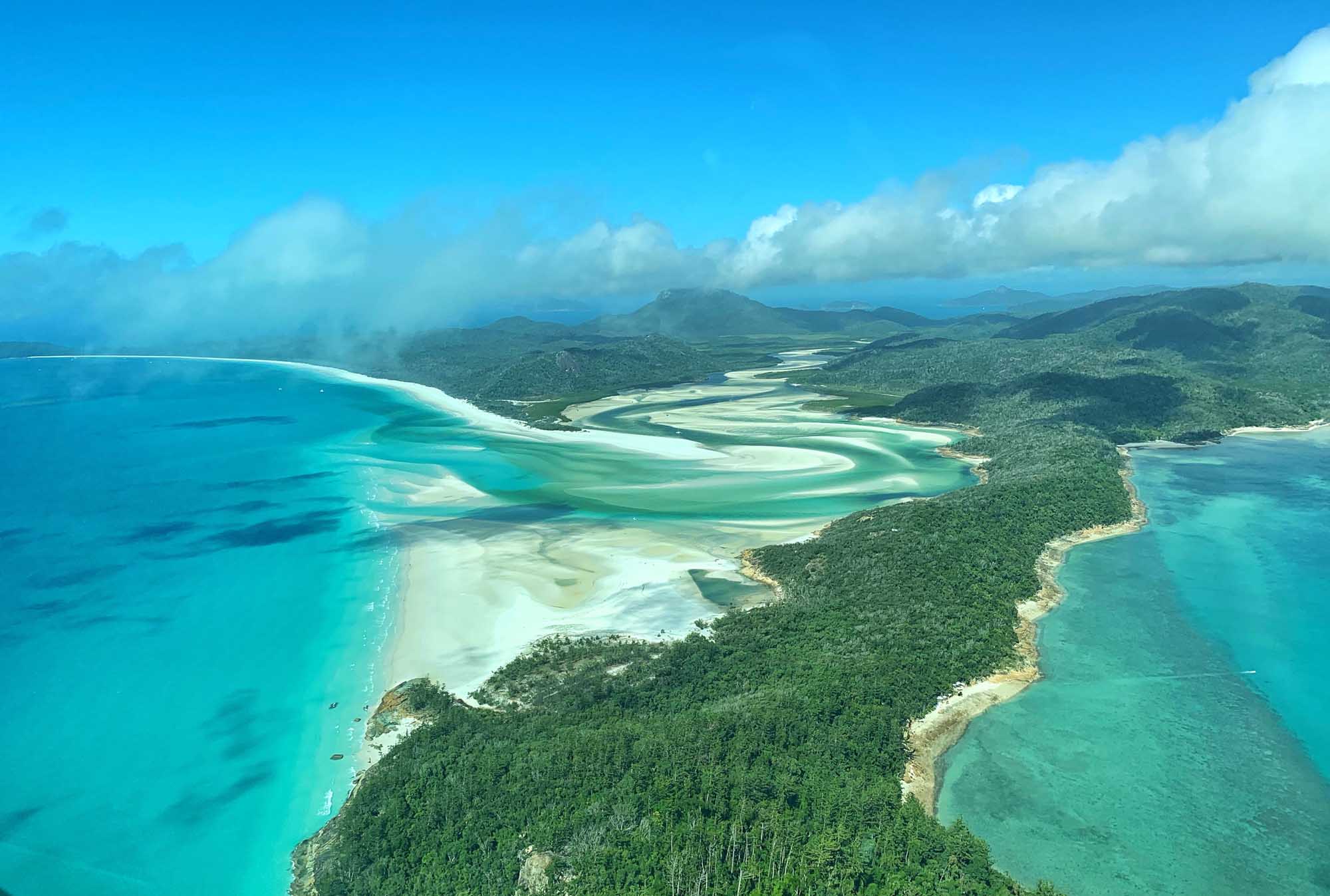 A stunning aerial photograph of the Whitsunday Islands, Australia, showcasing pristine turquoise waters, white sand beaches, and lush rainforest-covered islands with mountains in the background under a bright blue sky with white clouds.