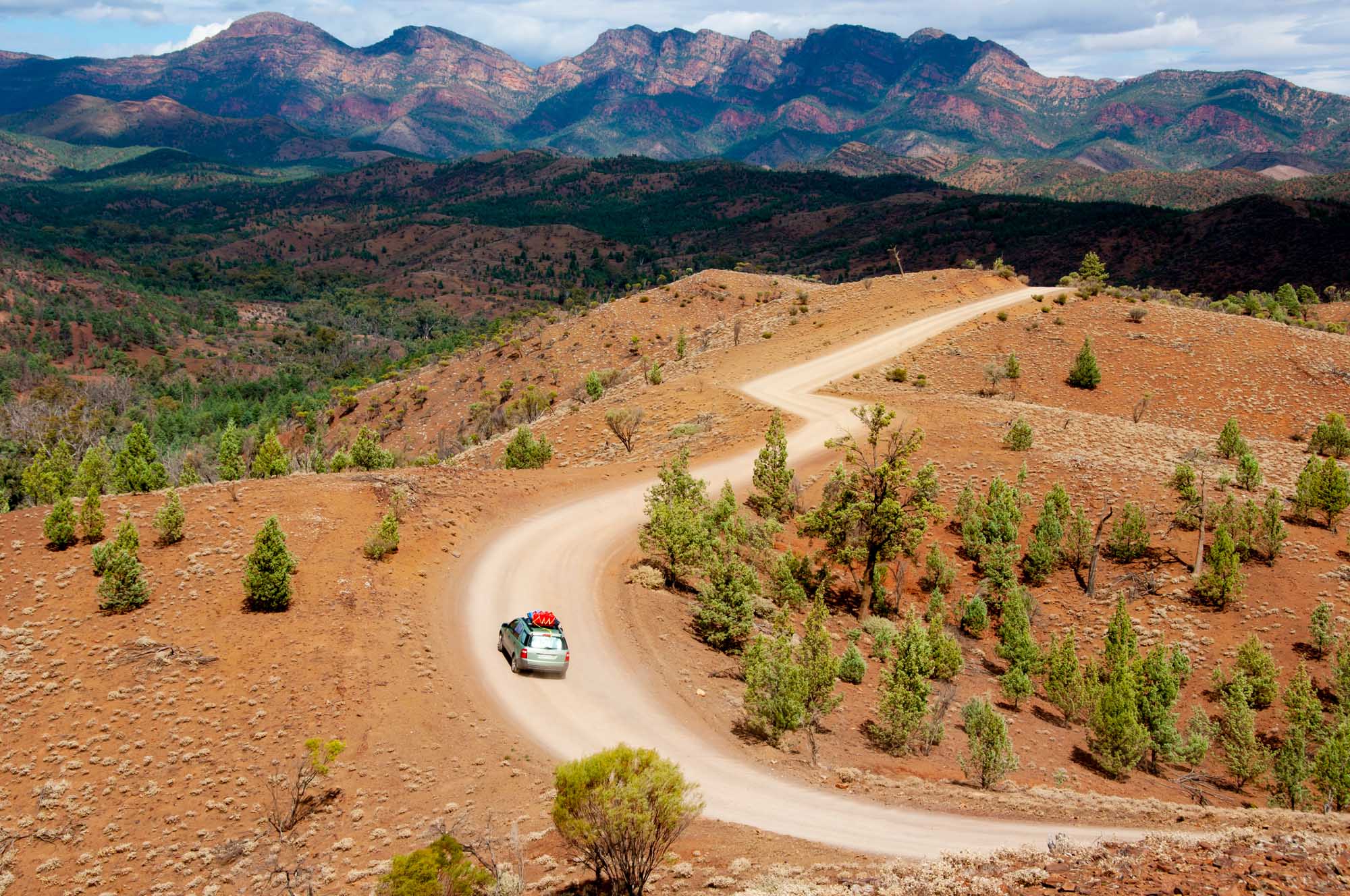A scenic aerial view of a winding dirt road cutting through the rugged terrain of Ikara-Flinders Ranges National Park in South Australia, with distinctive colored mountain ridges in the background and sparse native vegetation dotting the landscape.