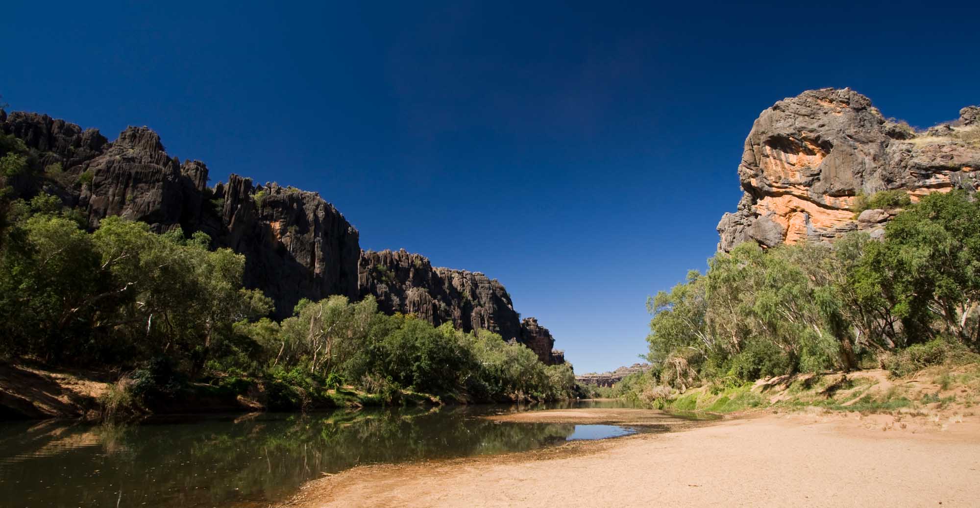 A stunning view of Windjana Gorge in Western Australia's remote Kimberley region, featuring towering sandstone cliffs framing a calm freshwater river lined with lush green vegetation against a brilliant blue sky. The dramatic geological formations and serene waterway showcase one of Australia's most pristine natural landmarks.