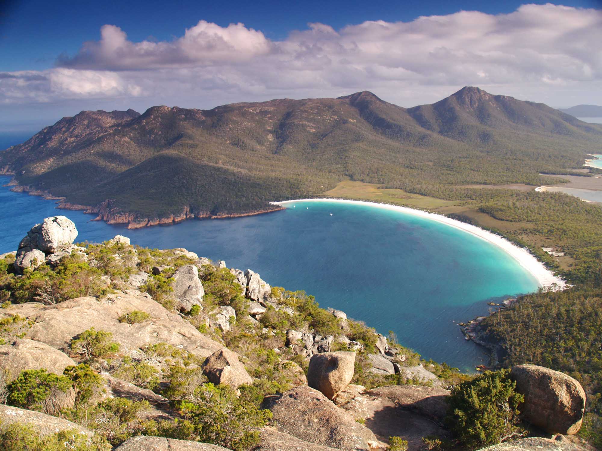 An aerial view of the stunning crescent-shaped Wineglass Bay at Freycinet National Park in Tasmania, featuring pristine turquoise waters, a white sand beach, and rugged granite peaks surrounded by native bushland. The image captures the dramatic coastal landscape from an elevated vantage point with rocky outcrops in the foreground.