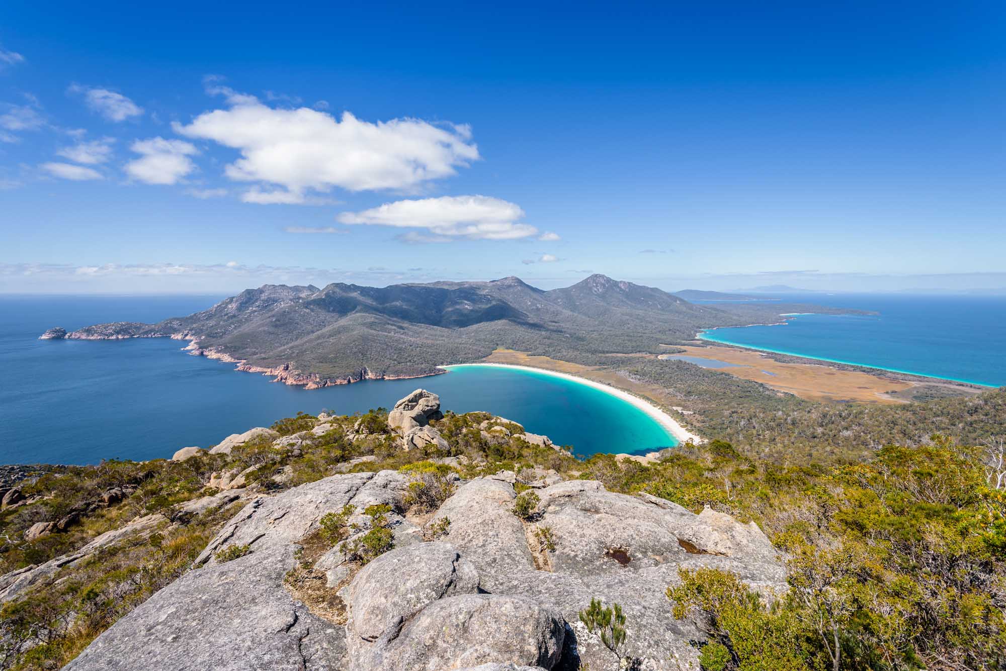 A stunning aerial view of Wineglass Bay in Freycinet National Park, Tasmania, featuring the iconic crescent-shaped white sand beach and turquoise waters surrounded by granite mountains and eucalyptus forest, photographed from the rocky summit of Mount Amos.