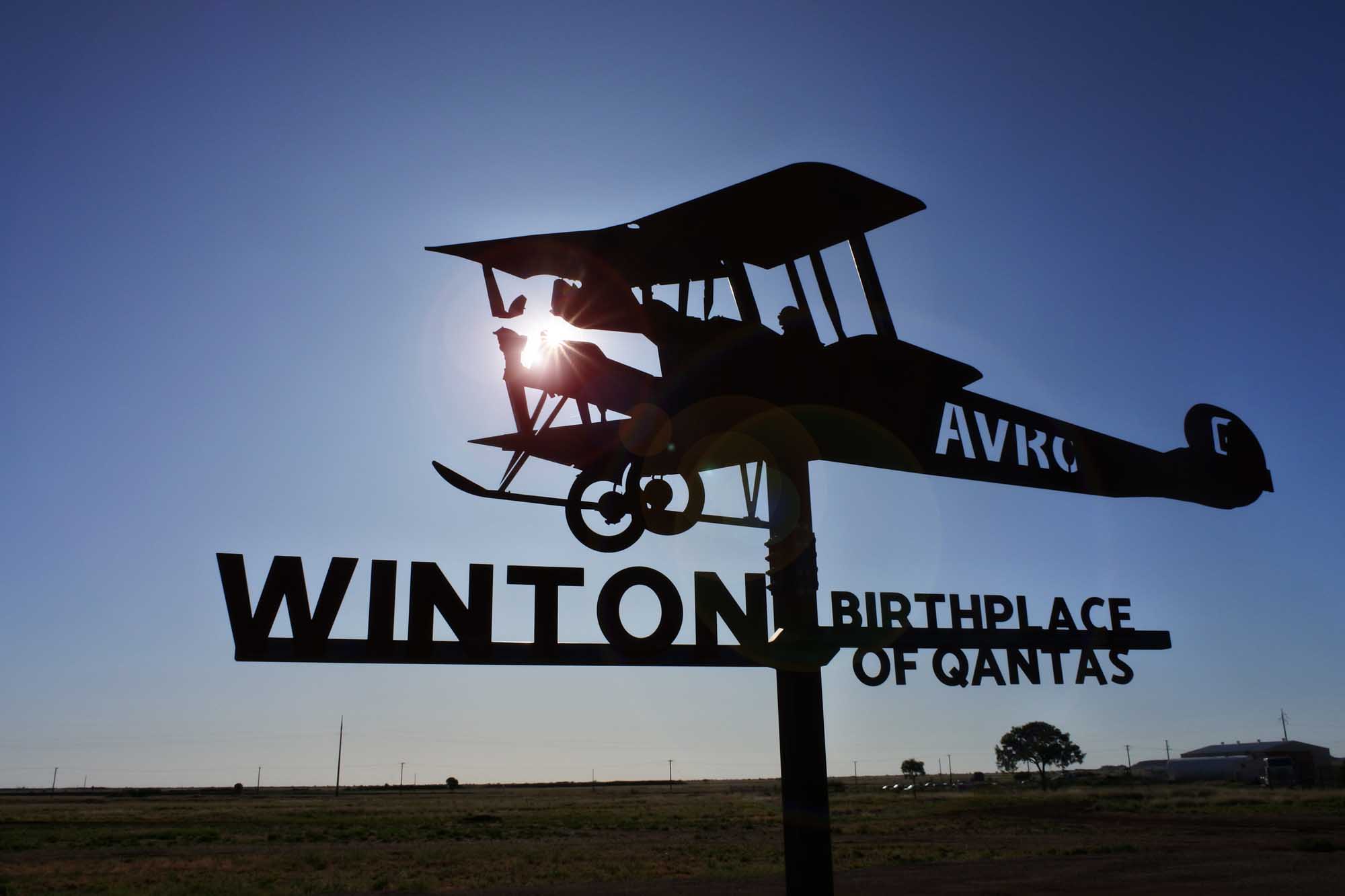 A silhouetted metal sculpture of a vintage biplane mounted on a pole against a blue sky, with 'WINTON BIRTHPLACE OF QANTAS' text displayed below. The sign commemorates Winton, Queensland, where Qantas airline was founded in 1920, set against a flat, rural Australian landscape.