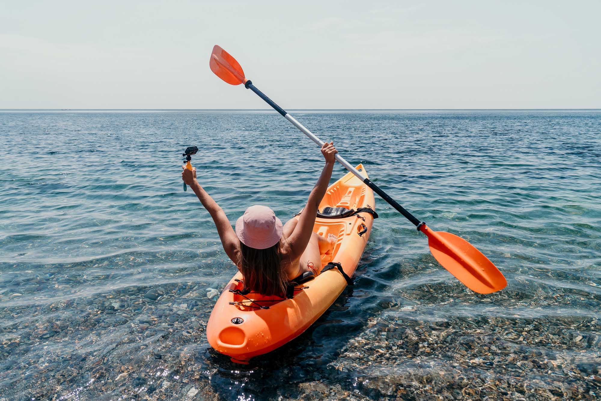 A woman paddles an orange kayak in shallow, clear turquoise ocean water near the shore, raising her