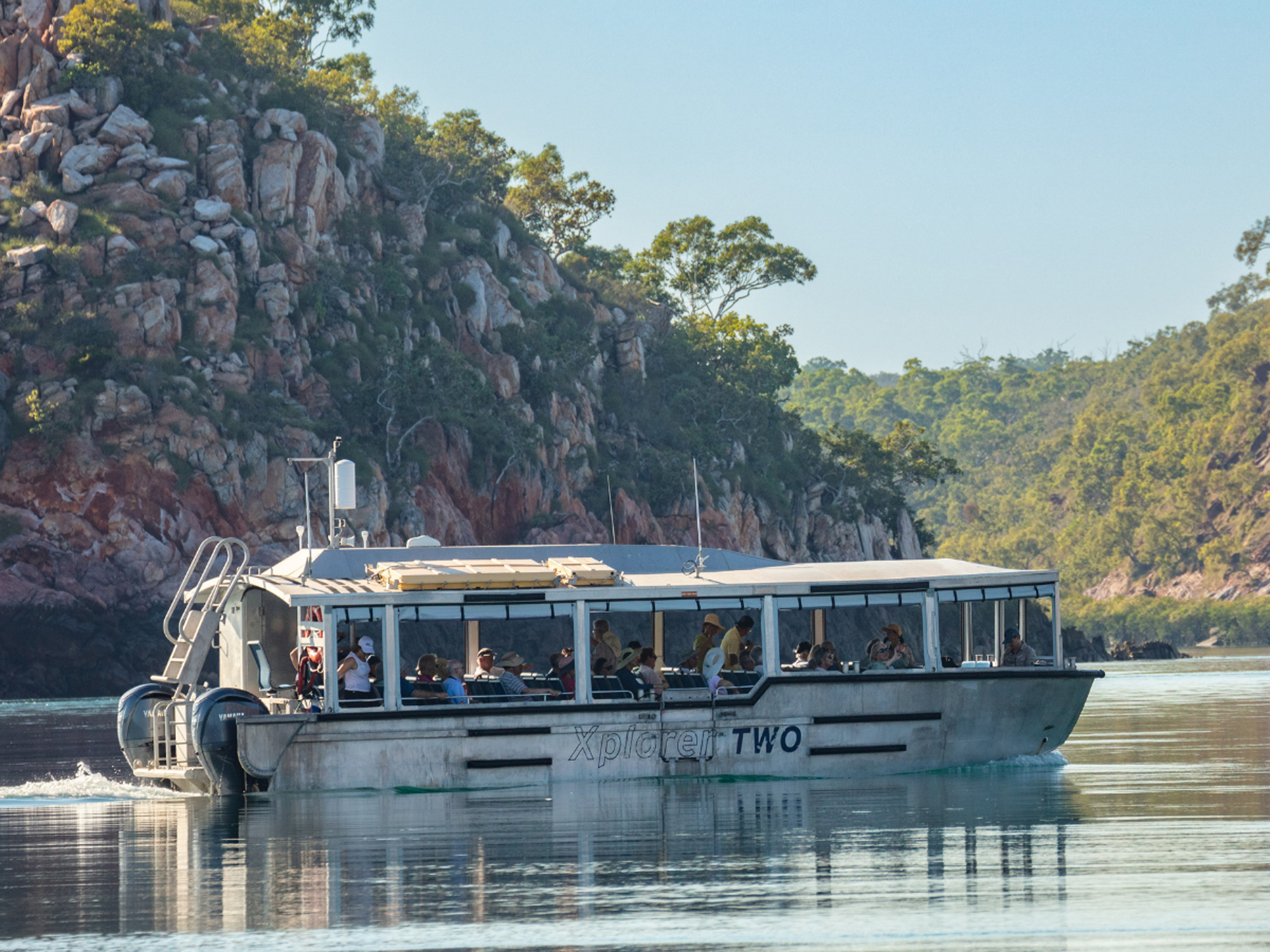 A scenic river cruise vessel named Xplorer Two navigates through a pristine waterway flanked by dramatic red rock cliffs and lush vegetation. The modern glass-enclosed passenger cabin is filled with tourists enjoying the natural landscape under clear skies.