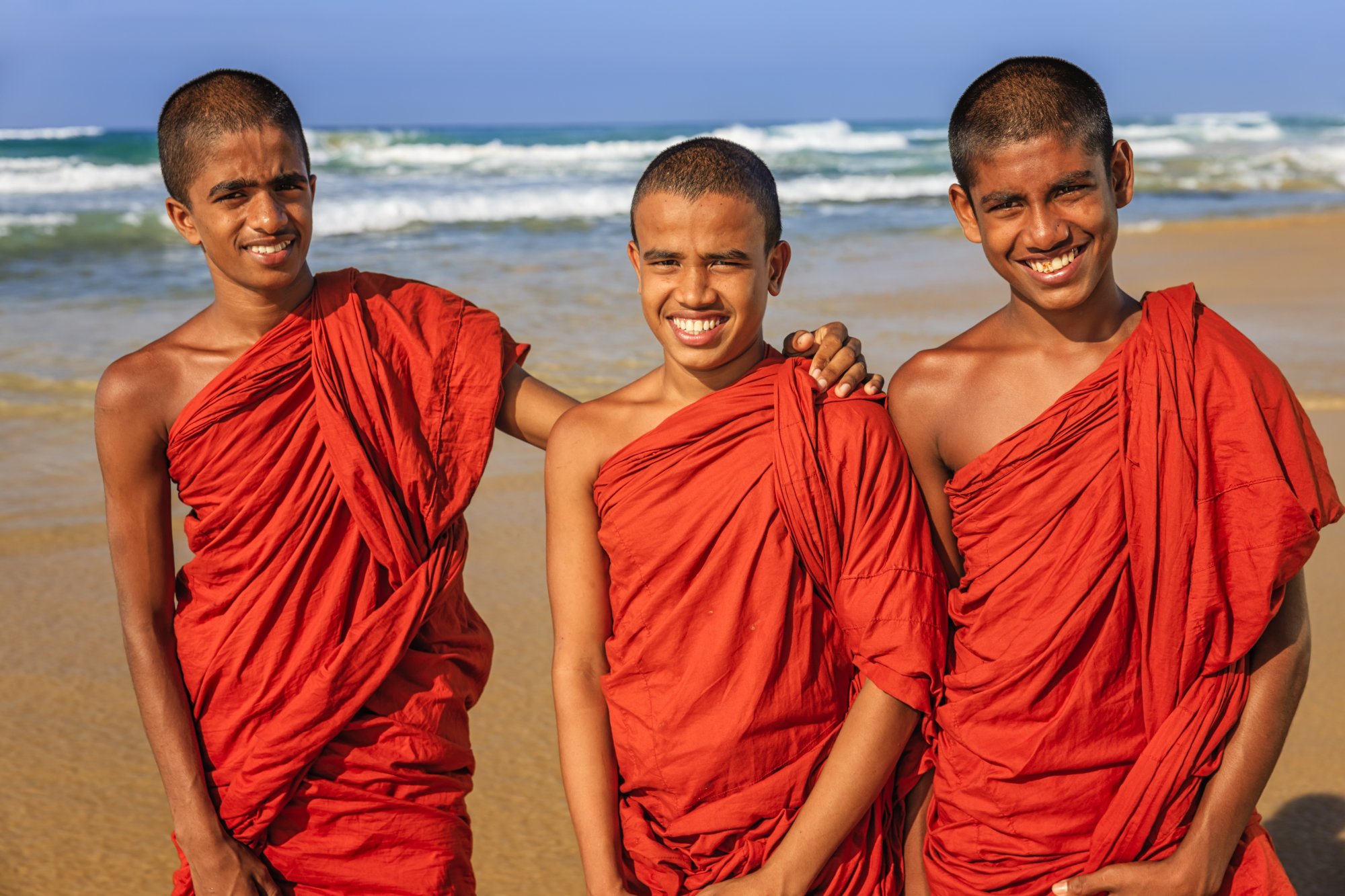 Three young Buddhist monks wearing traditional red robes stand together on a sandy beach with ocean