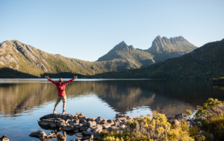 A hiker with arms raised standing on lakeside rocks facing jagged mountain peaks reflected in still water