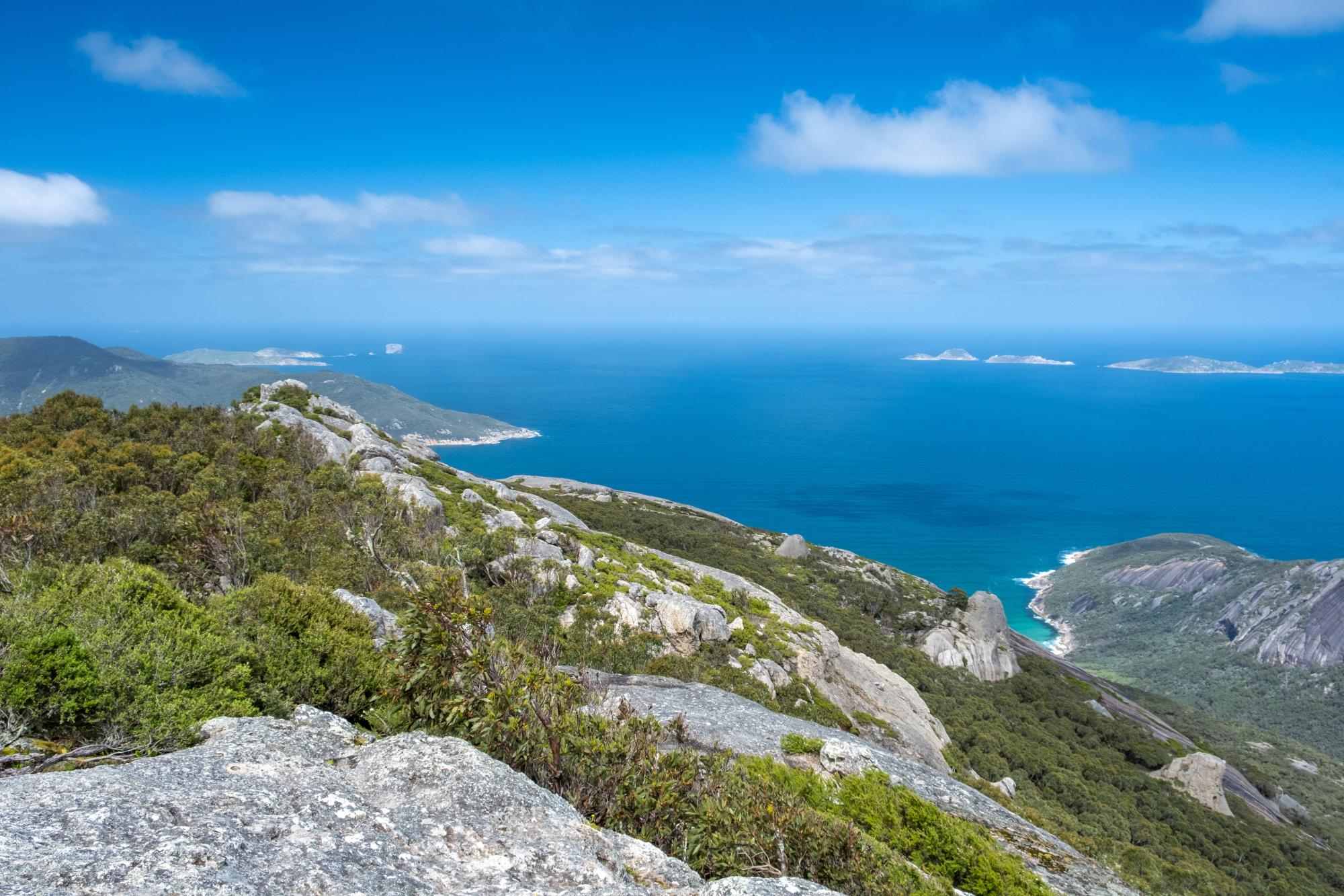 A sweeping coastal view from a granite mountain summit looking out over a deep blue sea dotted with small rocky islands.