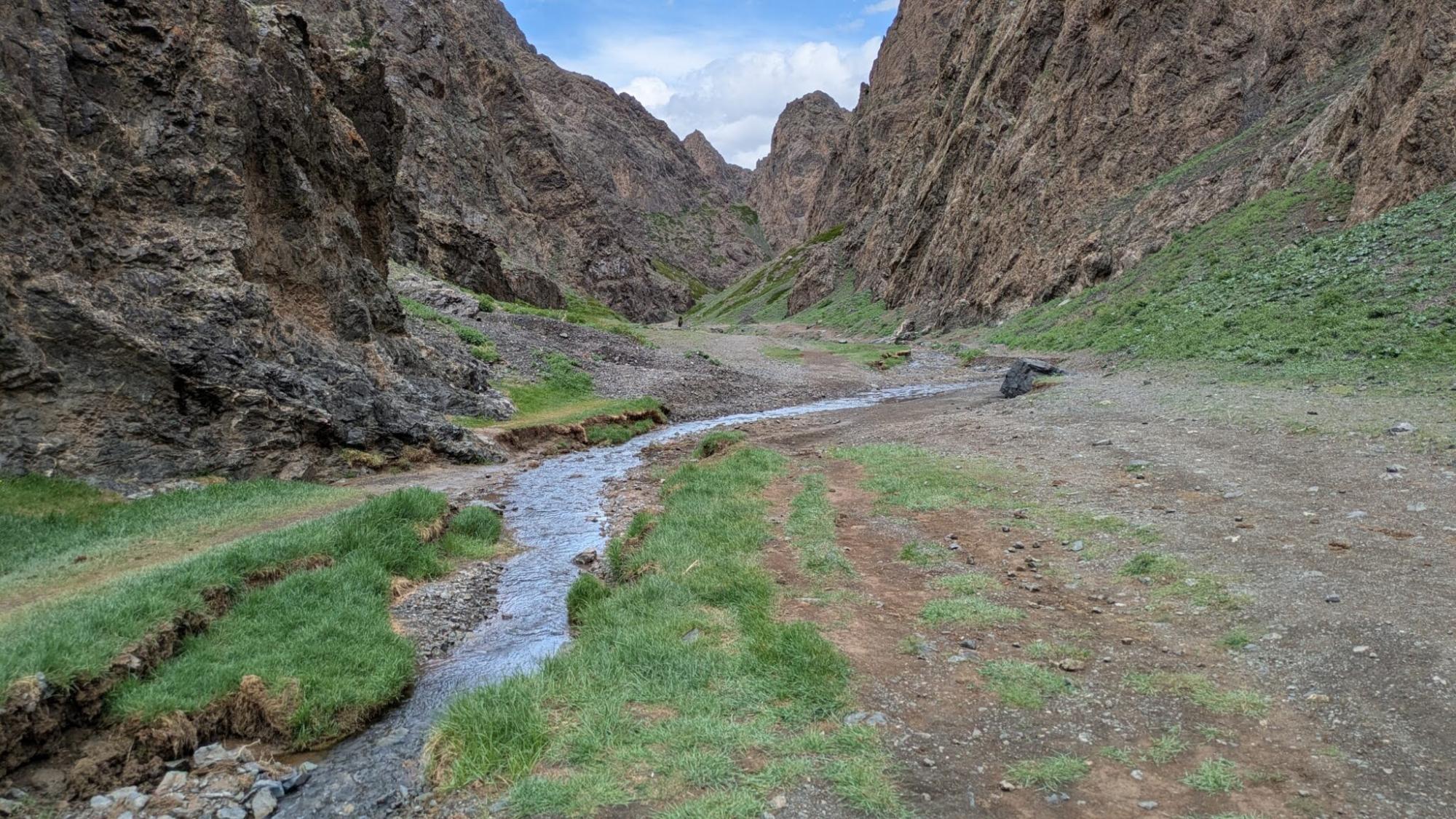 A dramatic mountain canyon with steep rocky cliffs on both sides, a small blue stream running