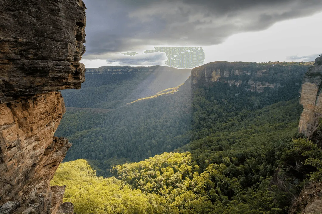 Sunlight beams through clouds over a dense green forest valley surrounded by steep rocky cliffs.